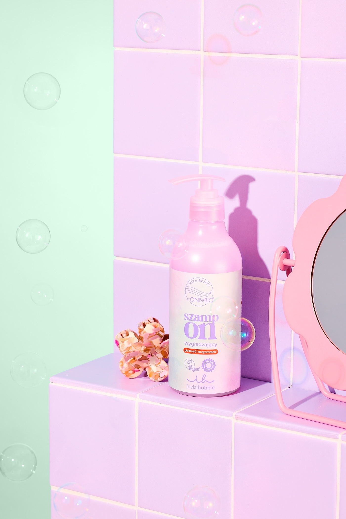 Szampon wygładzający o rozplątującej formule OnlyBio x invisibobble, w tle fioletowe płytki łazienkowe i lusterko w różowej oprawie.
