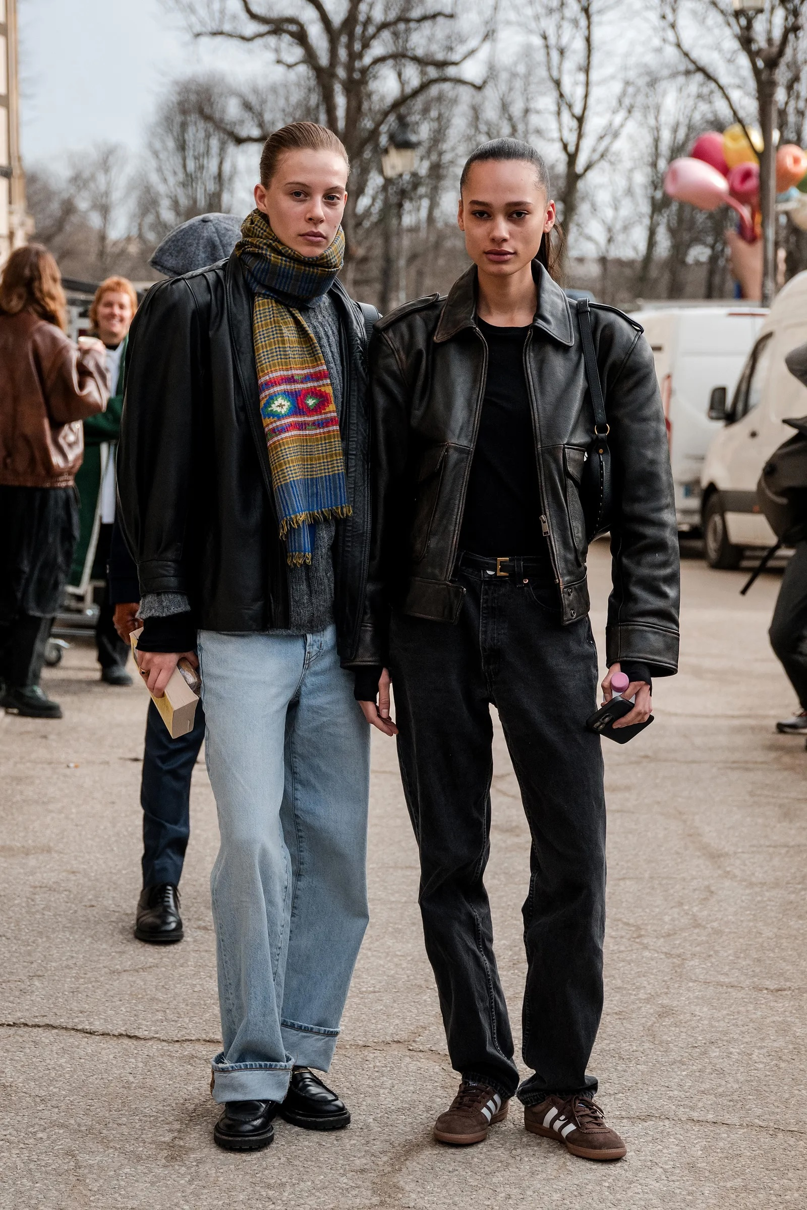 Dwie modelki stoją w jeansach, jedna w brązowych sneakersach Adidas Samba, druga w czarnej skórzanej kurtce.