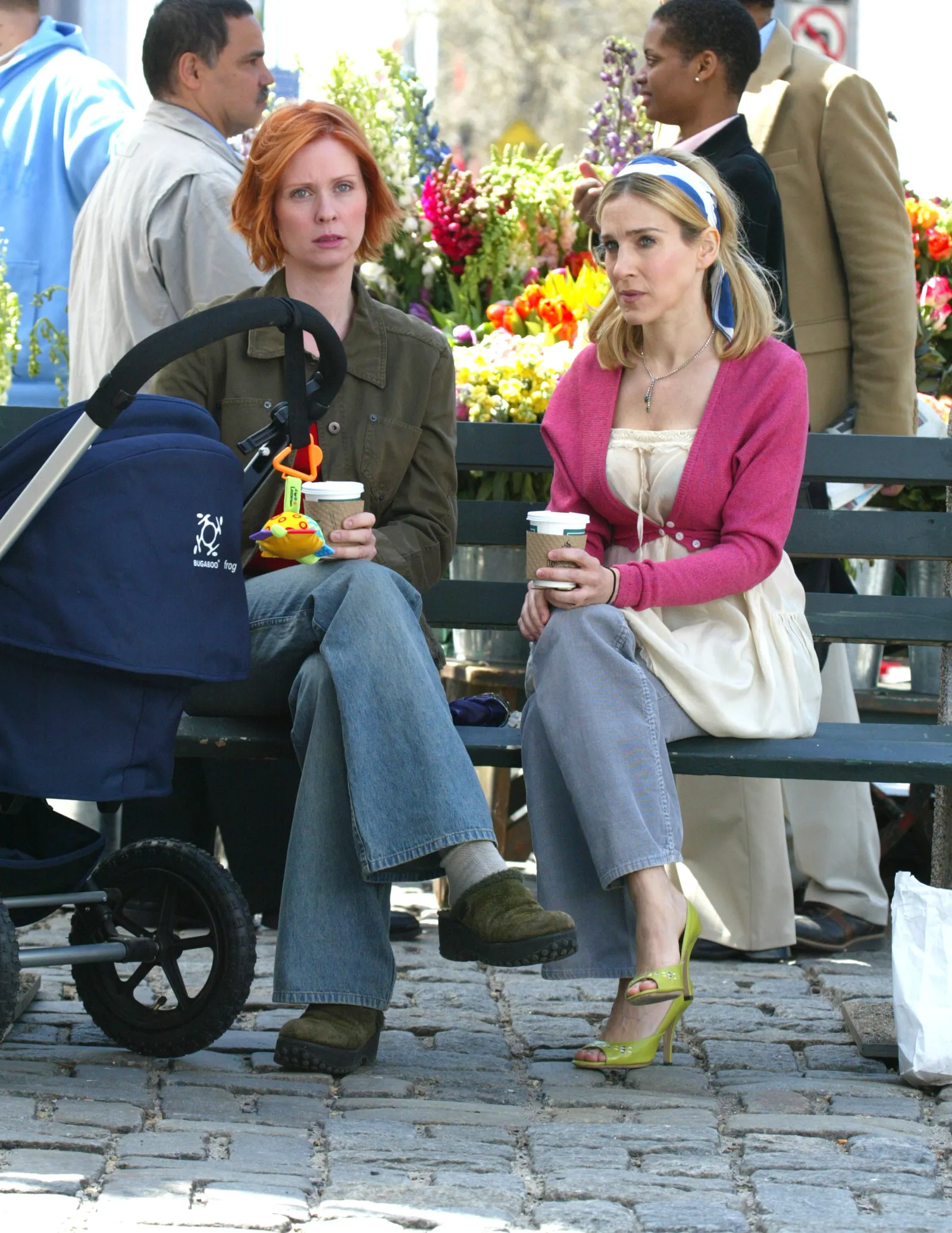 Cynthia Nixon jako Miranda Hobbes i Sarah Jessica Parker jako Carrie Bradshaw siedzą na ławce z kawą w otoczeniu kwiatów.