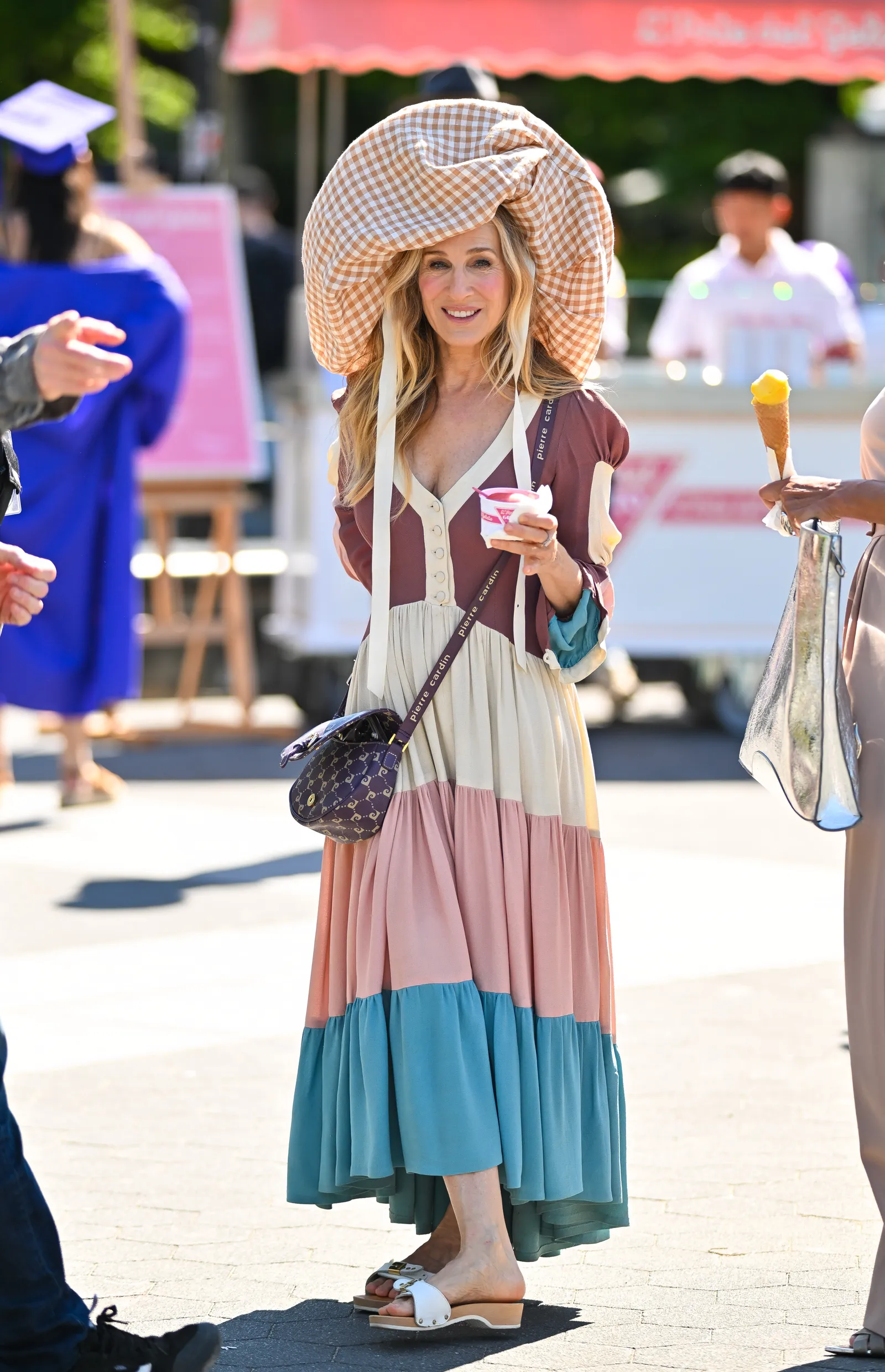 Sarah Jessica Parker jako Carrie Bradshaw w kolorowej sukience i dużym kapeluszu, trzymająca jedzenie.