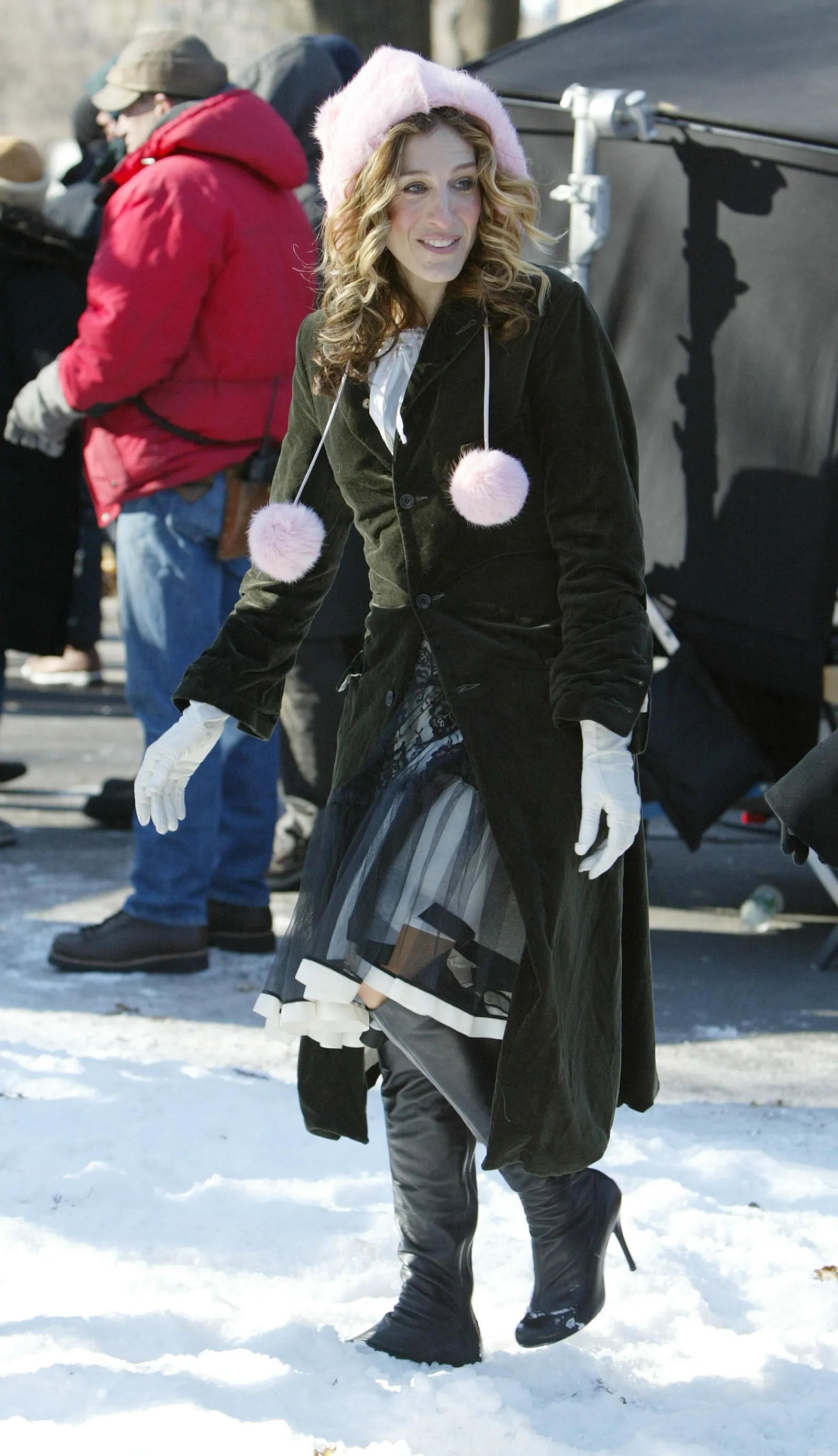Sarah Jessica Parker jako Carrie Bradshaw w płaszczu, różowej czapce i długich kozakach, spaceruje po zaśnieżonym terenie.