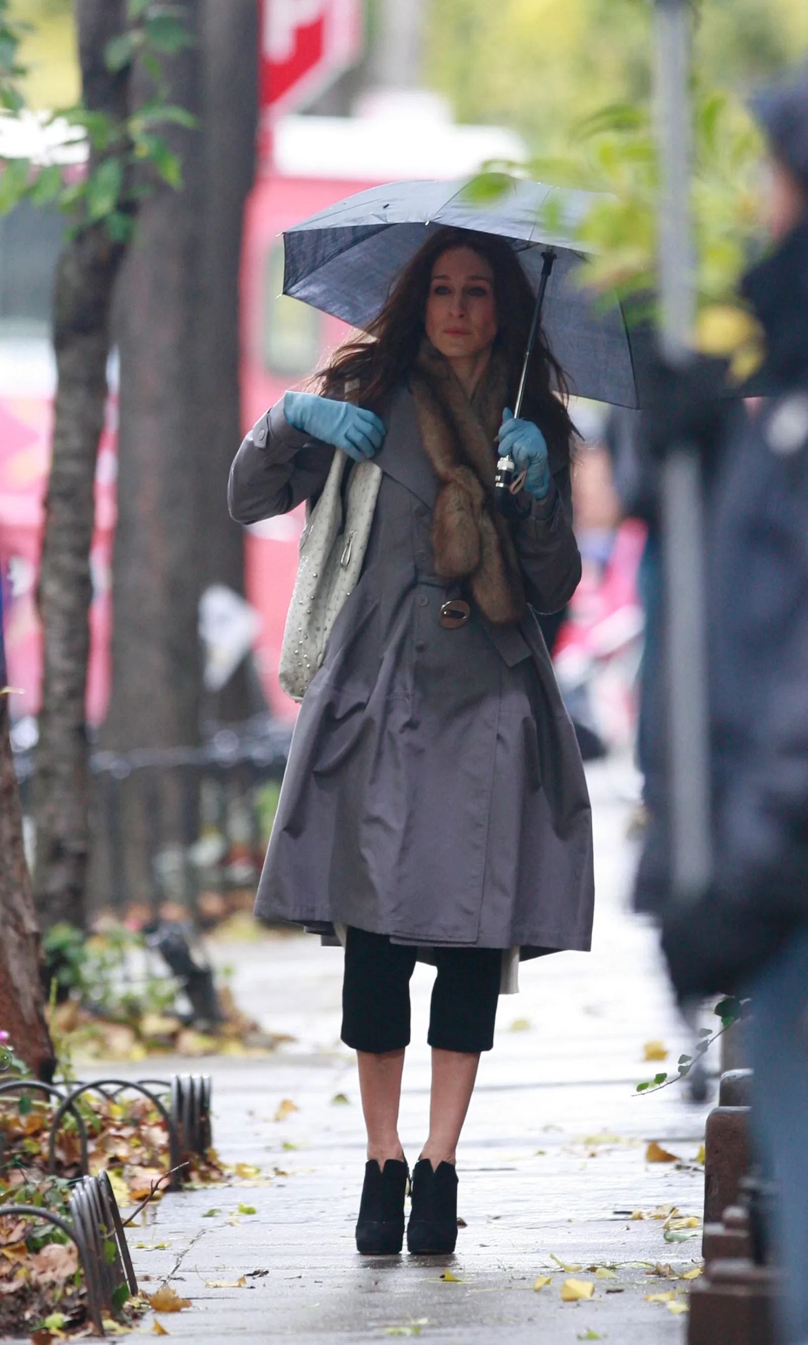 Sarah Jessica Parker jako Carrie Bradshaw, idąca ulicą w szarym płaszczu, z parasolką i rękawiczkami.