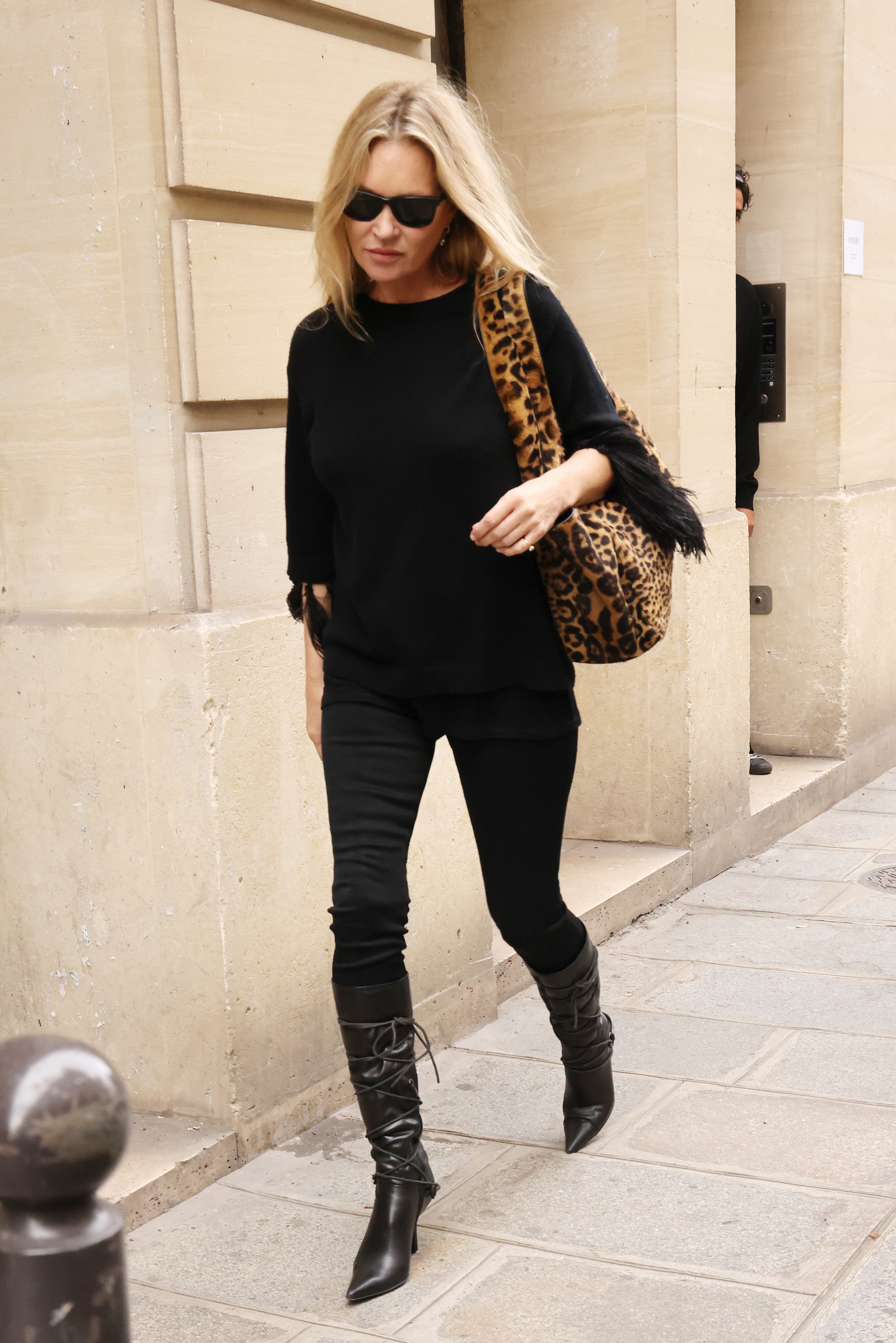 Kate Moss w czarnych skinny jeans, czarnych kozakach na szpilce oraz torebce w panterkę, przechadza się ulicą.