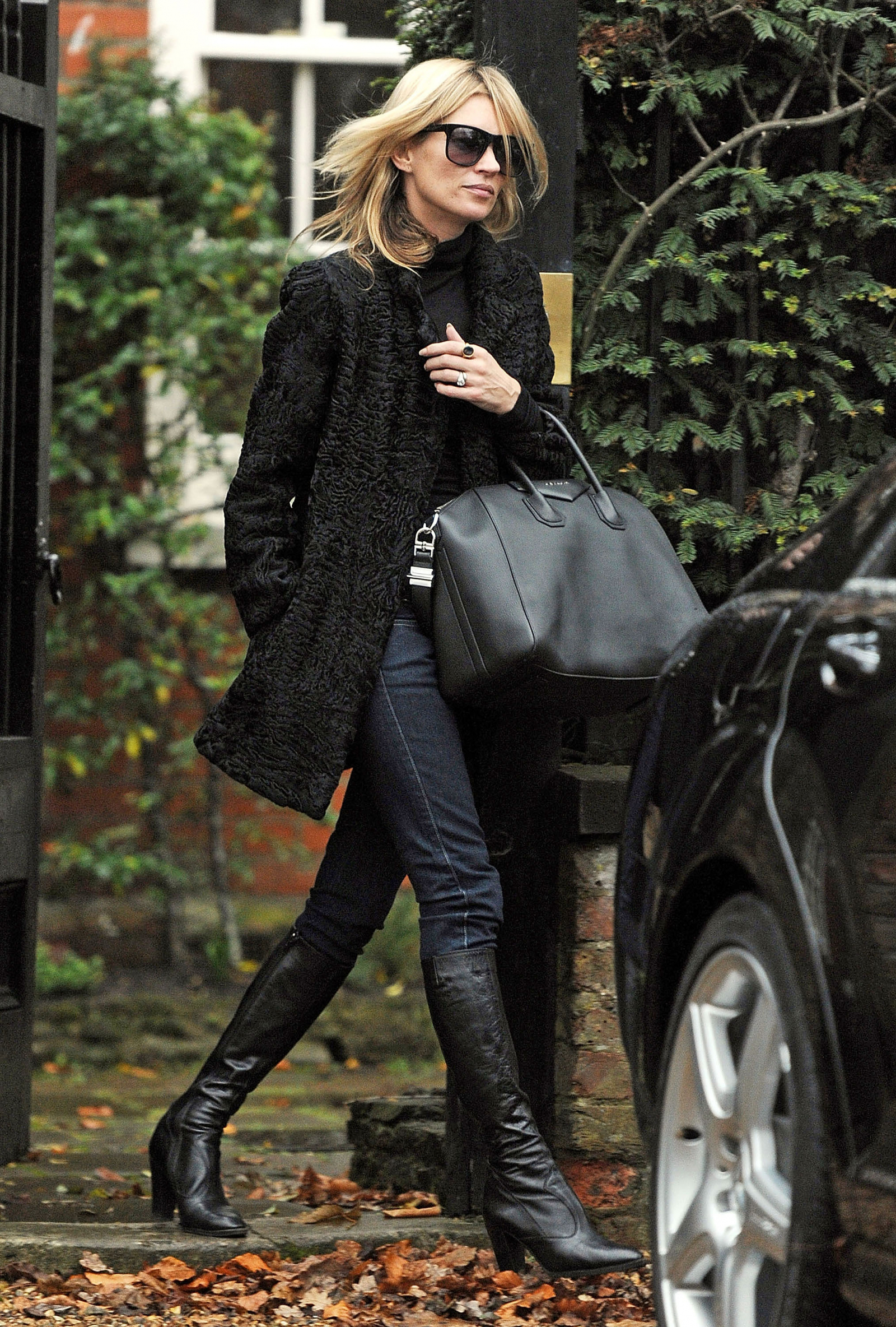Kate Moss w czarnym płaszczu, ciemne spodnie typu skinny oraz czarne, dopasowane kozaki, spaceruje z torbą.