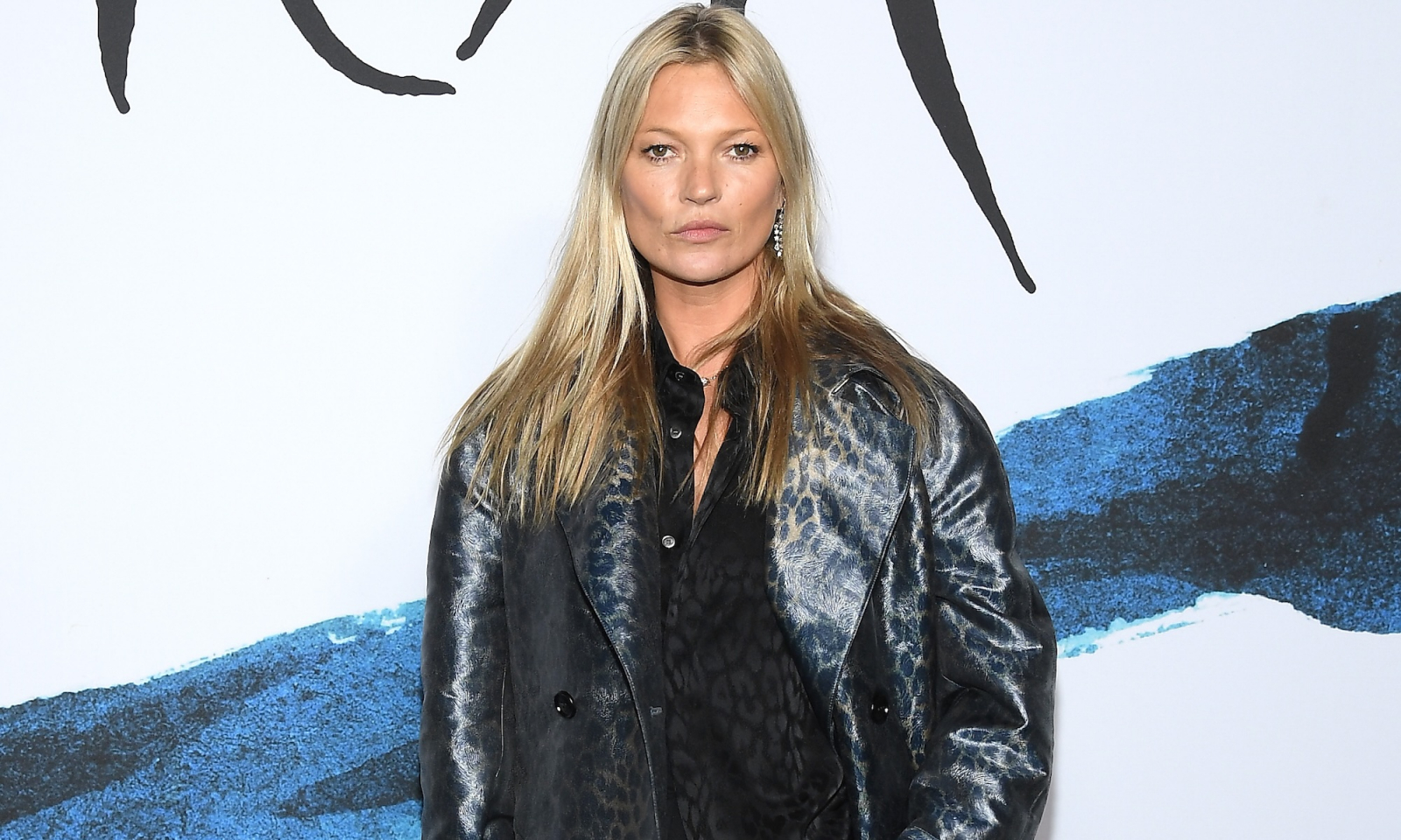 Supermodelka Kate Moss w eleganckim, połyskującym płaszczu, stoi na tle artystycznego muralu.