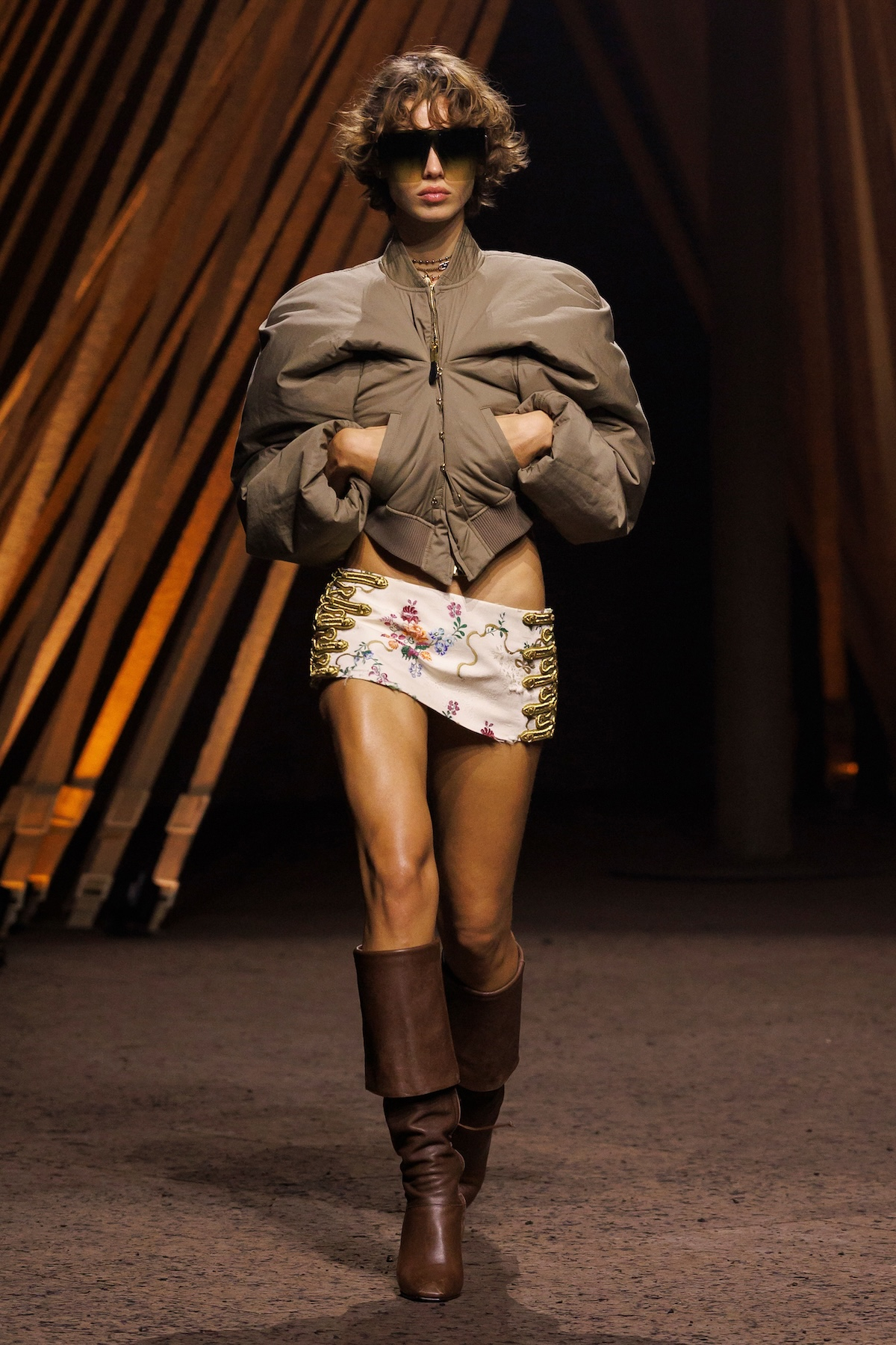 Modelka w krótkiej spódnicy i brązowych kozakach na obcasie, w oversize'owej kurtce, prezentuje modowy look na wybiegu.