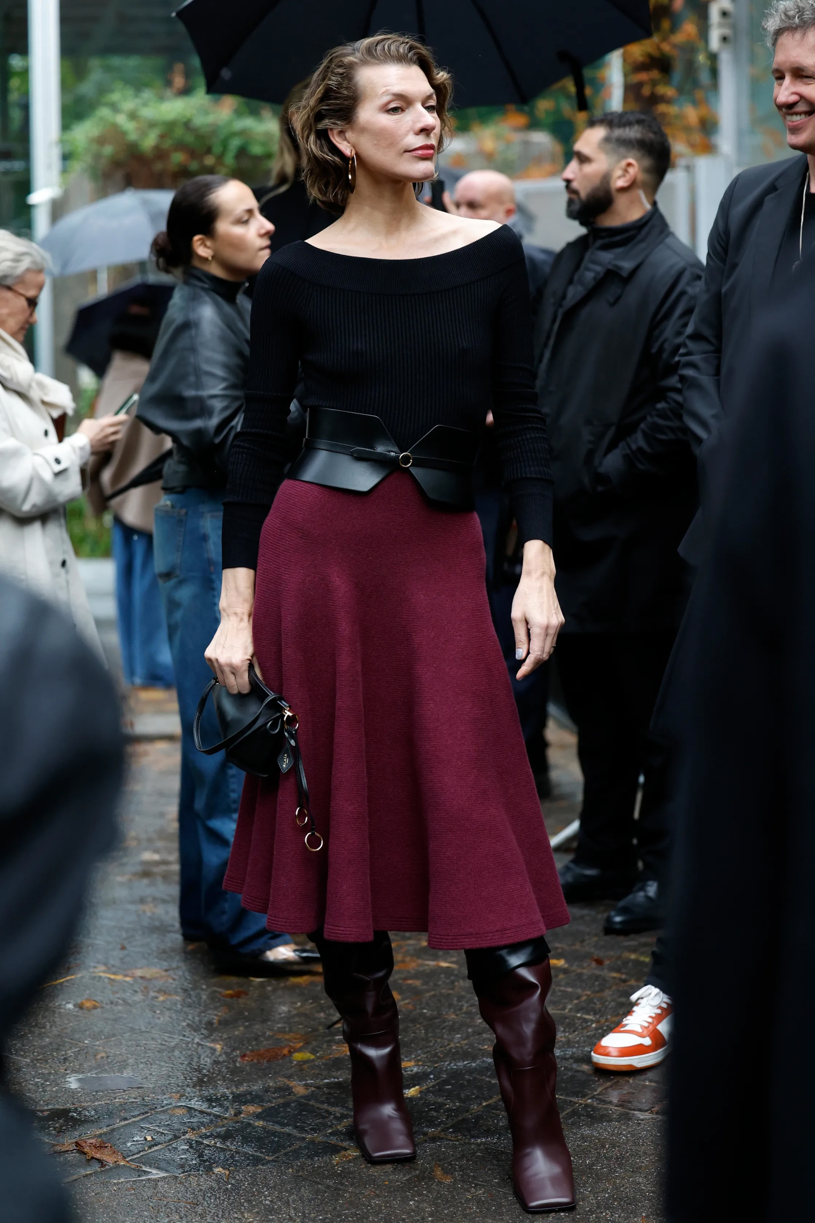 Milla Jovovich w czarnej bluzce, bordowej spódnicy midi i burgundowych kozaka z kwadratowym noskiem.