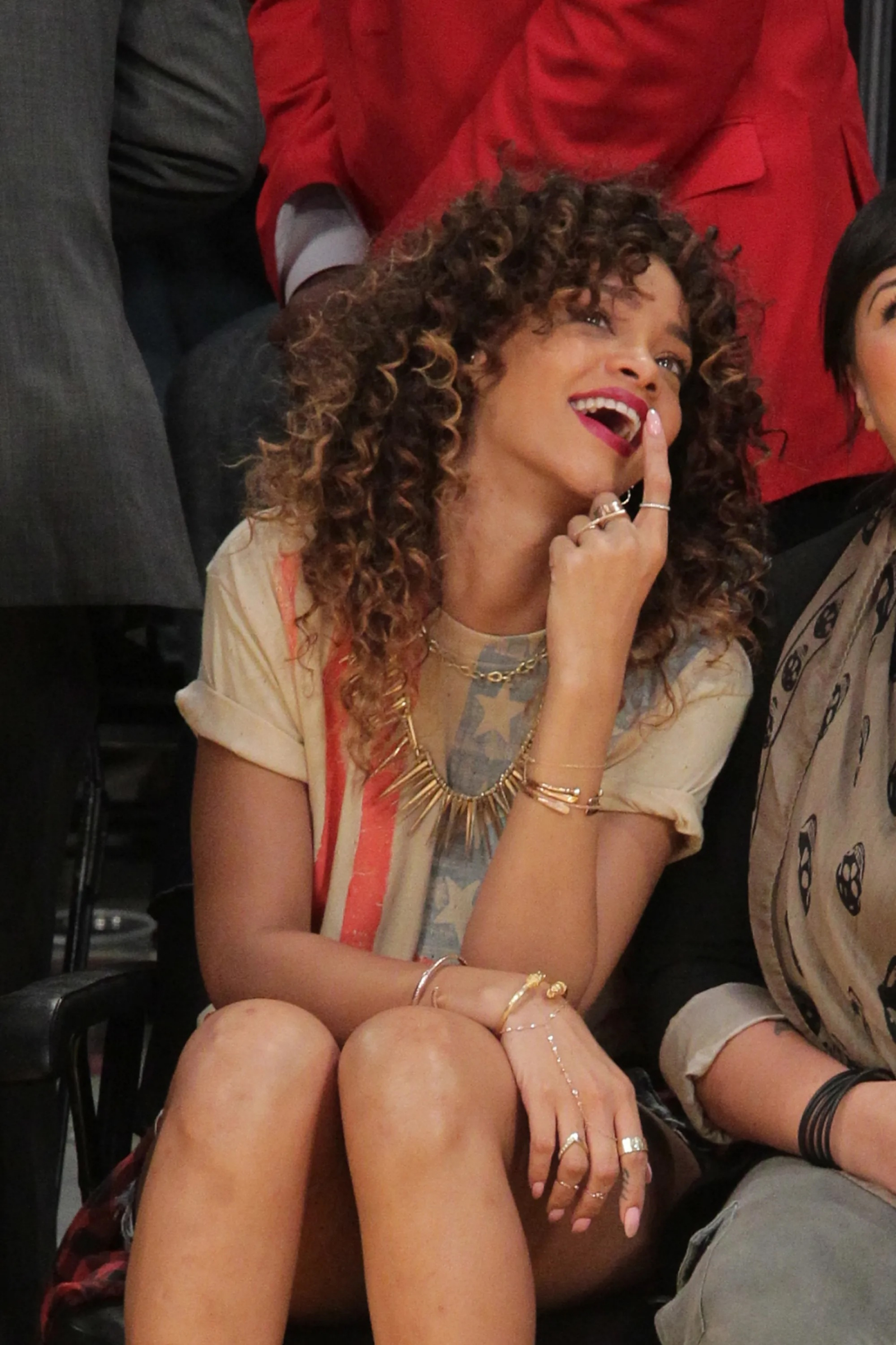 Rihanna w 2012 roku, z uśmiechem i złotą bransoletką na ręce, siedzi na trybunach.