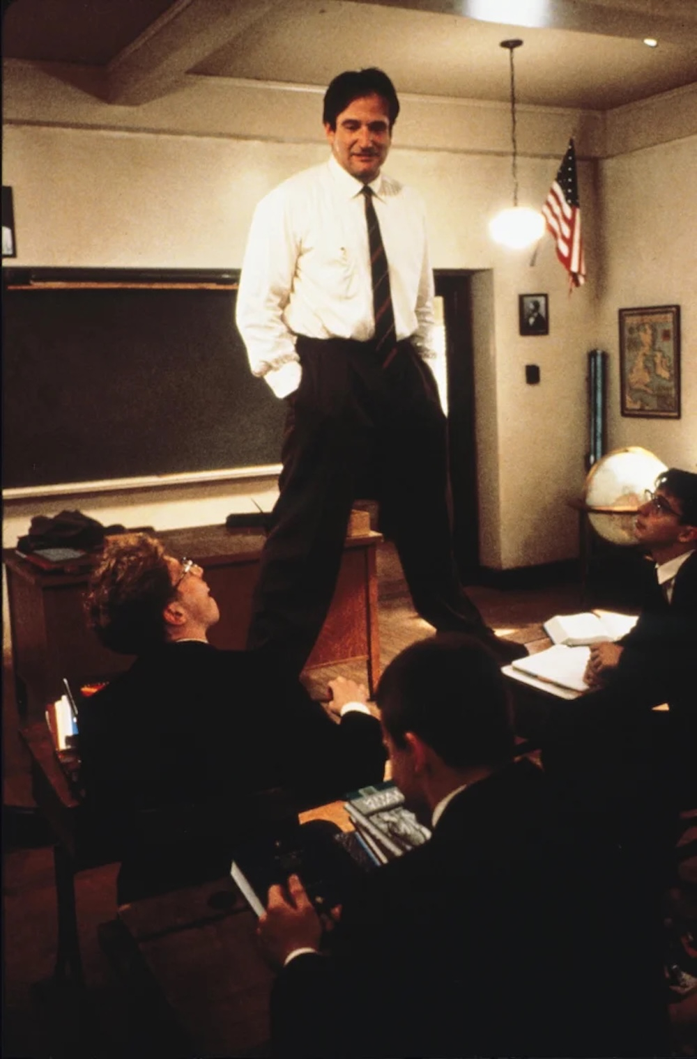 Robin Williams w filmie „Stowarzyszenie Umarłych Poetów” stoi na biurku, rozmawiając z uczniami w klasie.