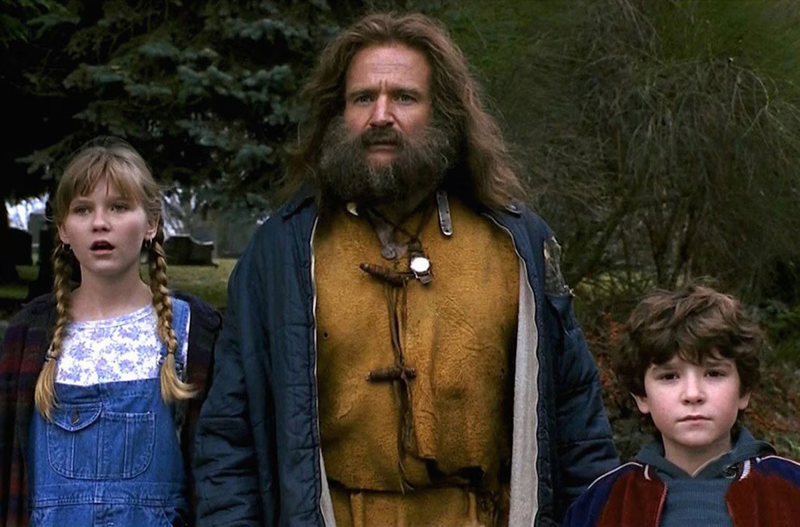 Robin Williams w filmie „Jumanji” stoi z dwojgiem dzieci, wyglądają zaskoczeni w scenerii leśnej.