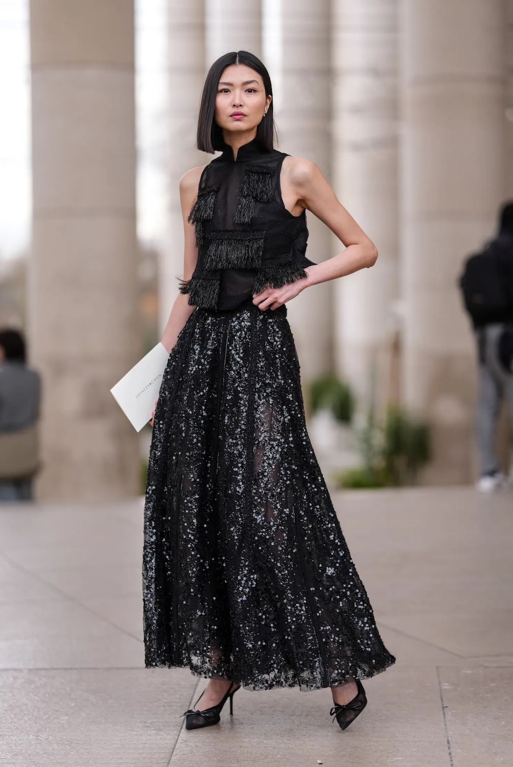 Modelka w długiej spódnicy z cekinami i bluzie z oryginalnym zdobieniem pozuje w eleganckiej scenerii.
