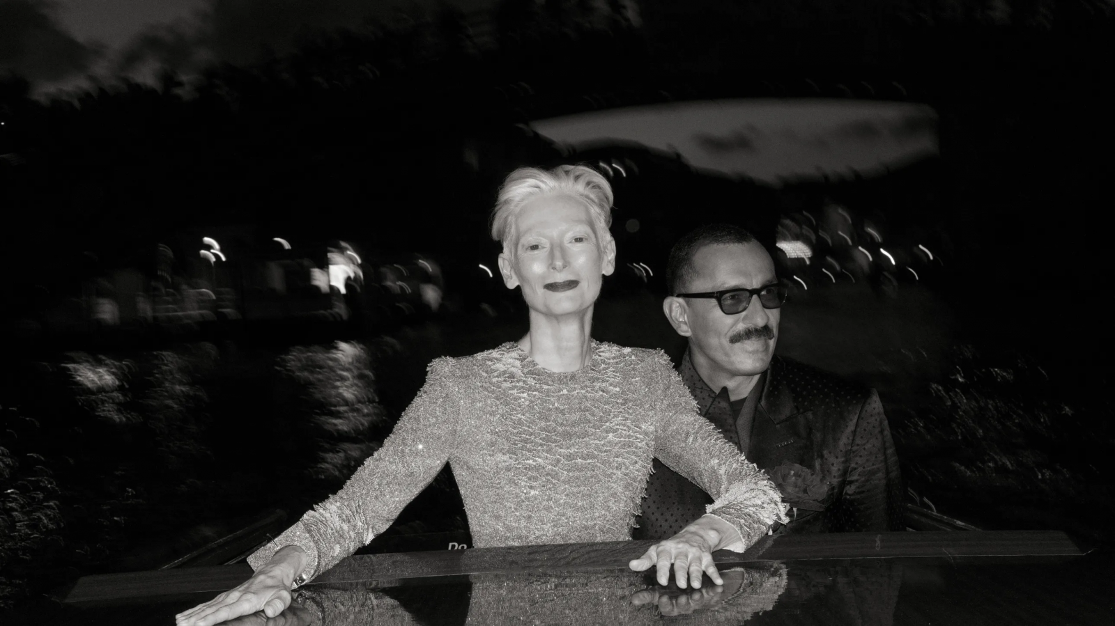 Tilda Swinton i Haider Ackermann podczas rejsu po Wenecji, ujęcie w monochromatycznym stylu.