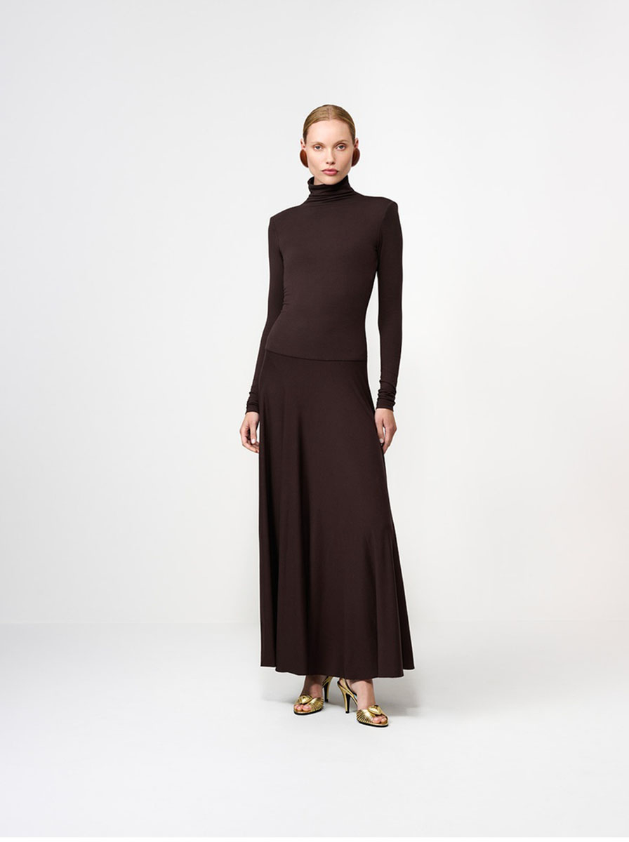 Modelka w brązowej sukience maxi z długim rękawem, stoi na białym tle, w eleganckich butach.