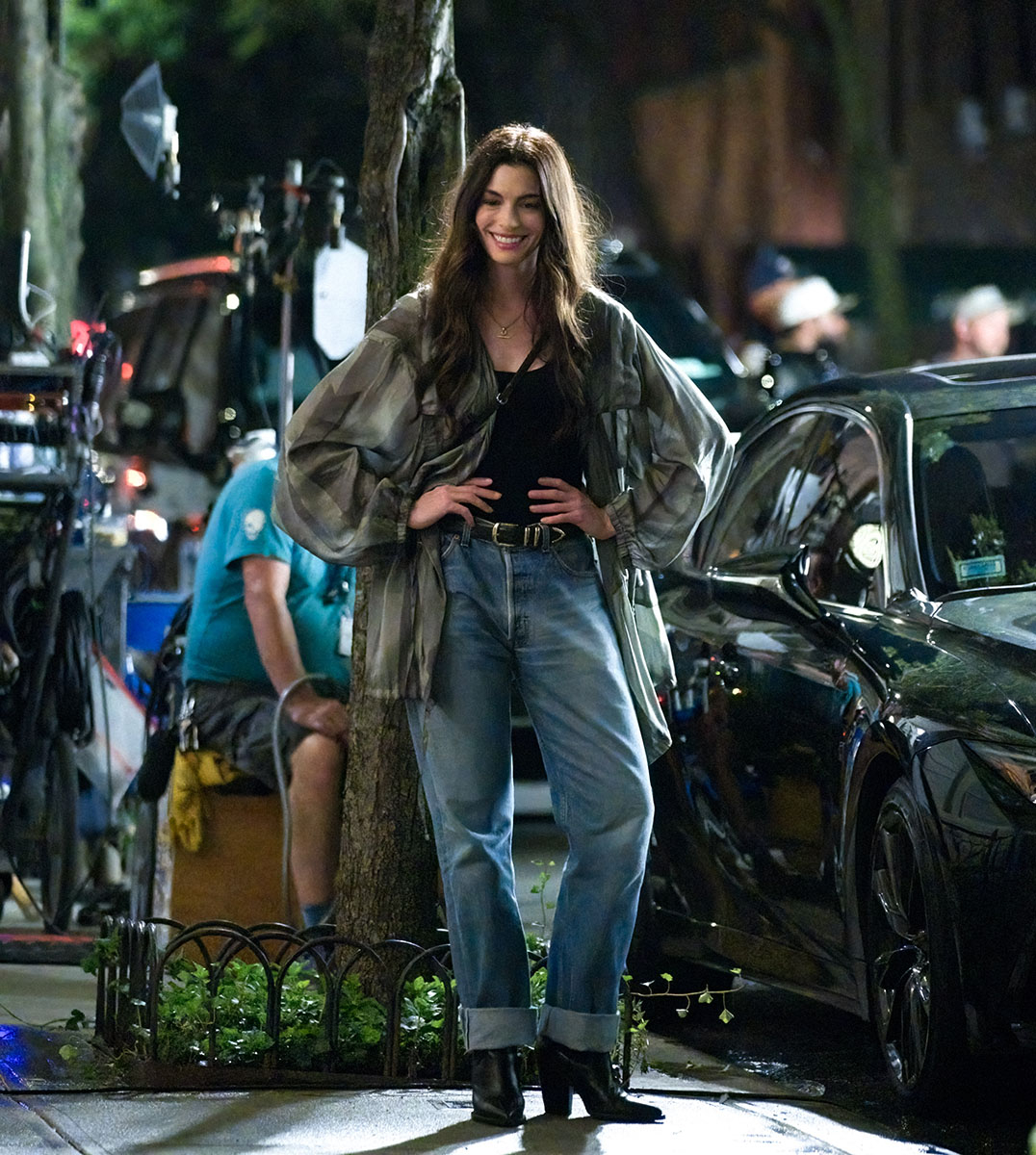 Anne Hathaway w luźnym stroju, uśmiechając się na planie zdjęciowym z widocznymi ludźmi w tle.