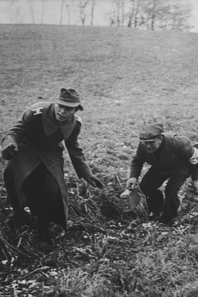 Żołnierze przeszukują teren, pochylając się nad ziemią, w otoczeniu zieleni.