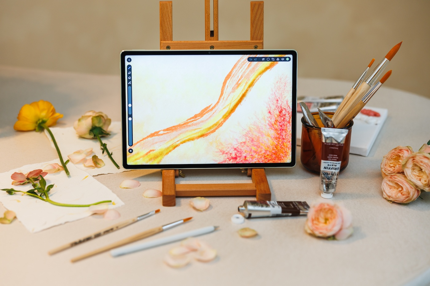 Na stole znajduje się tablet Huawei MatePad Pro z kolorowym rysunkiem, obok akcesoria: farby, pędzle i kwiaty.