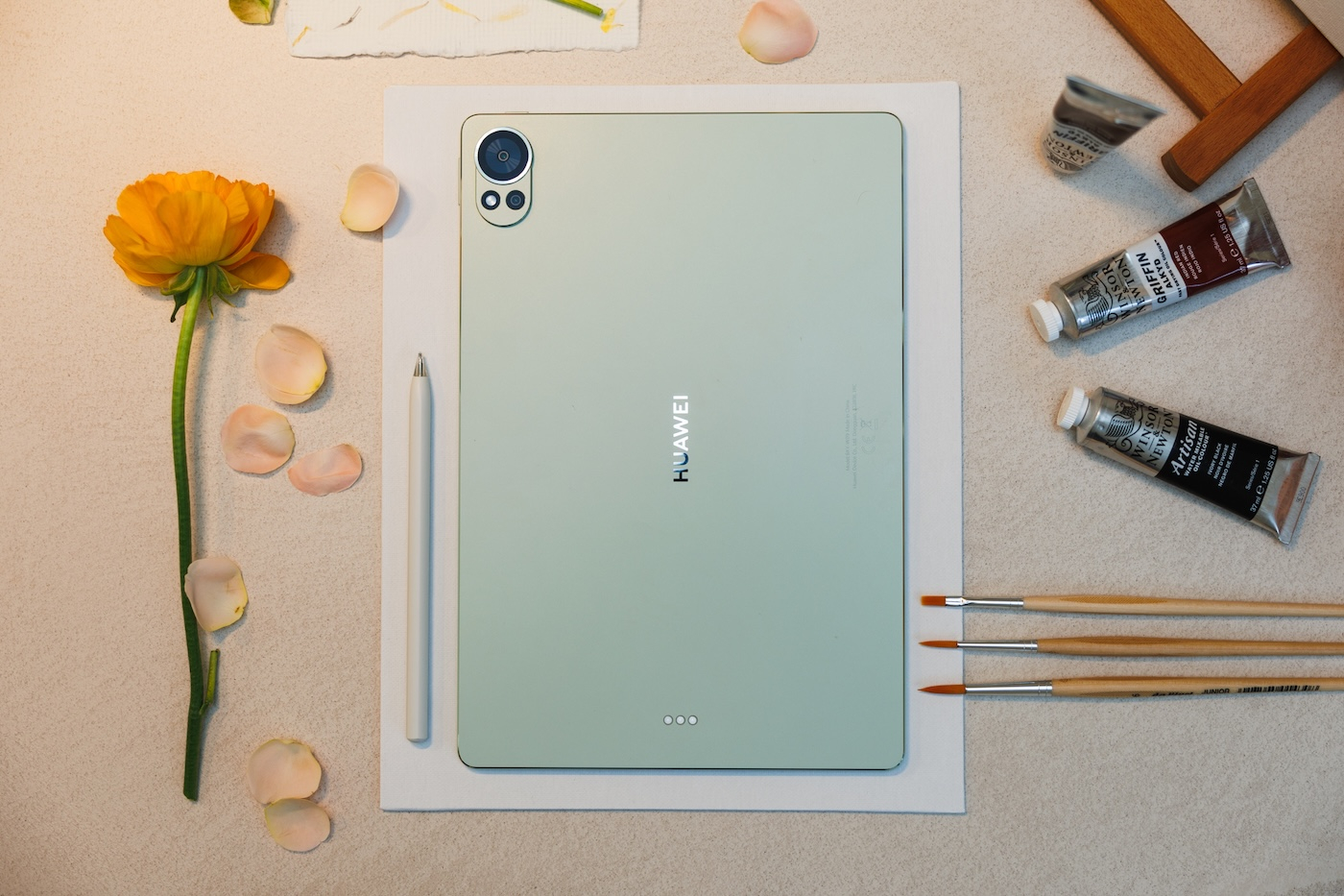 Tablet Huawei MatePad Pro leży na białej kartce, obok akcesoria: pędzle, farby i kwiat.