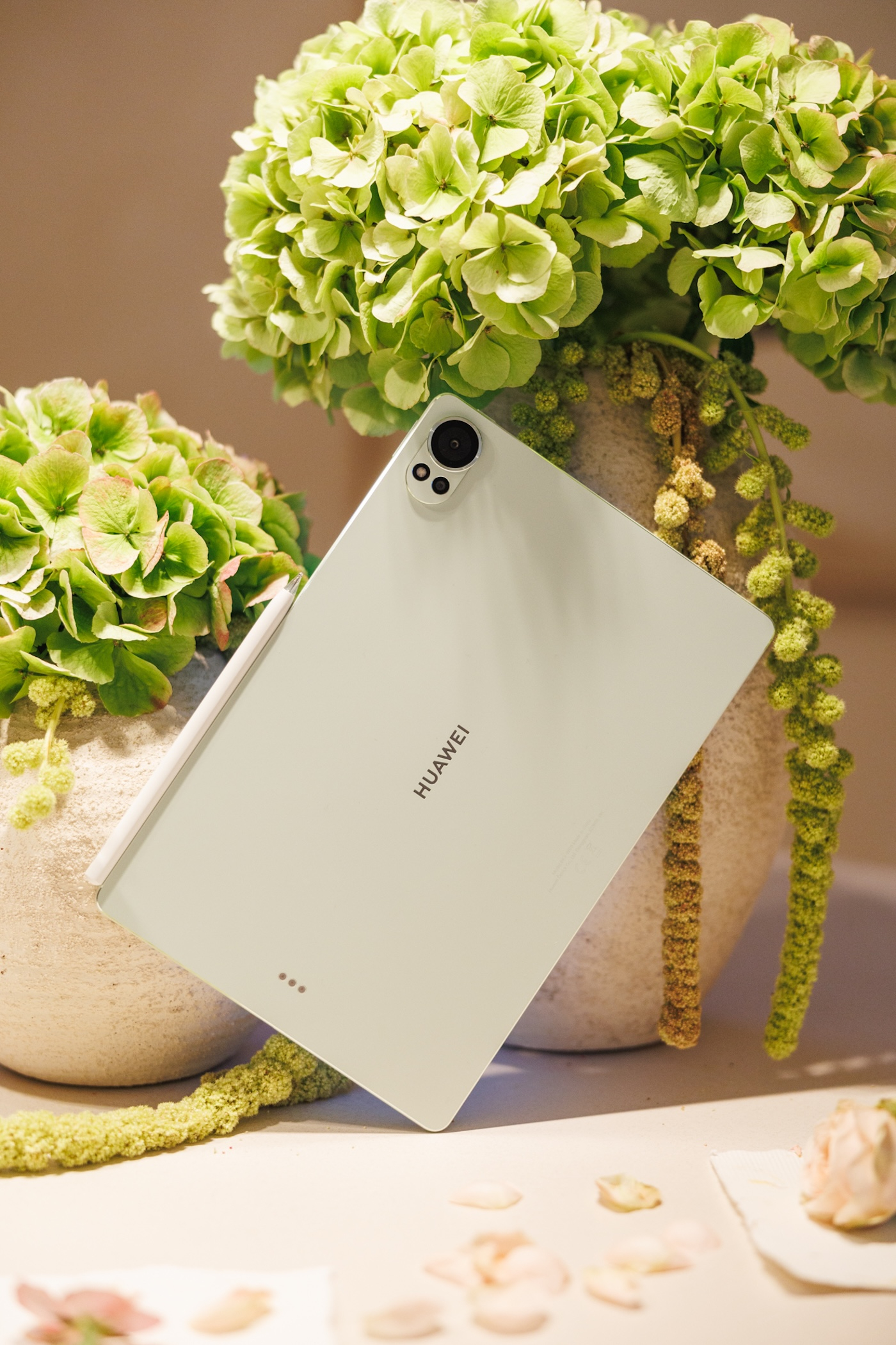 Tablet Huawei MatePad Pro oparty o doniczki z zielonymi kwiatami. Tło neutralne, z delikatnymi akcentami florystycznymi.