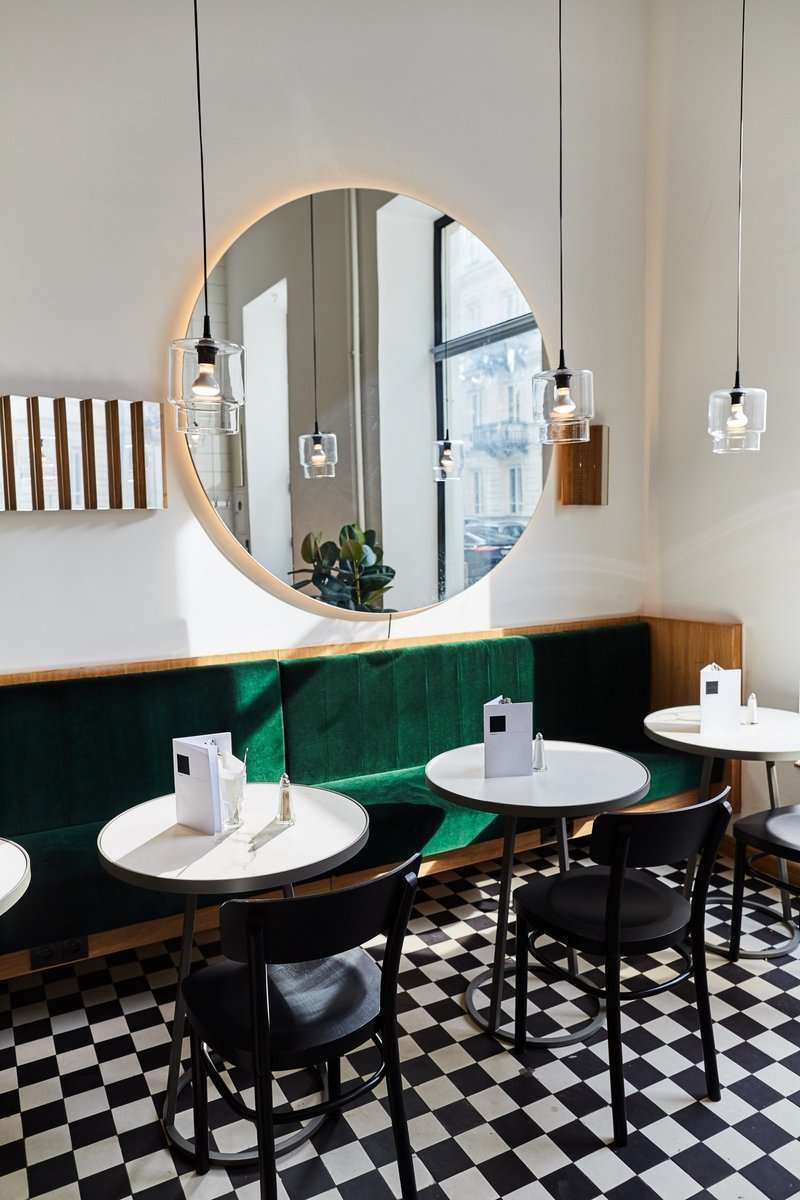 Wnętrze kawiarni Krem przedstawiające zielone, welurowe kanapy , okrągłe stoły i lustro w złotej ramie wiszące na ścianie nad kanapą.
