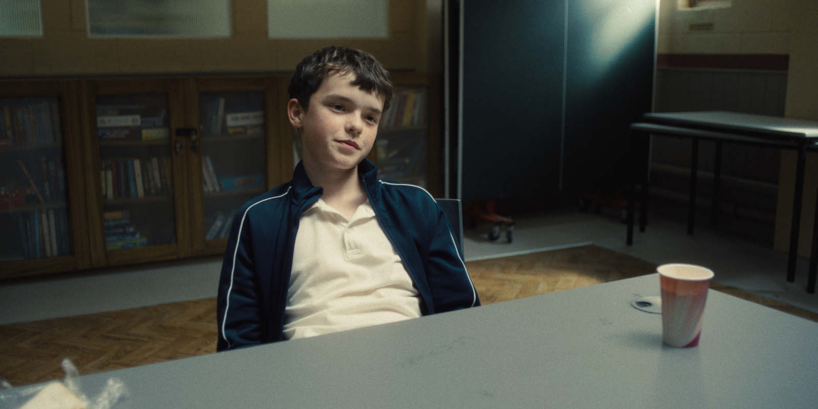 Owen Cooper w scenie z serialu Netflix „Dojrzewanie”, siedzi przy stole w pomieszczeniu z książkami.