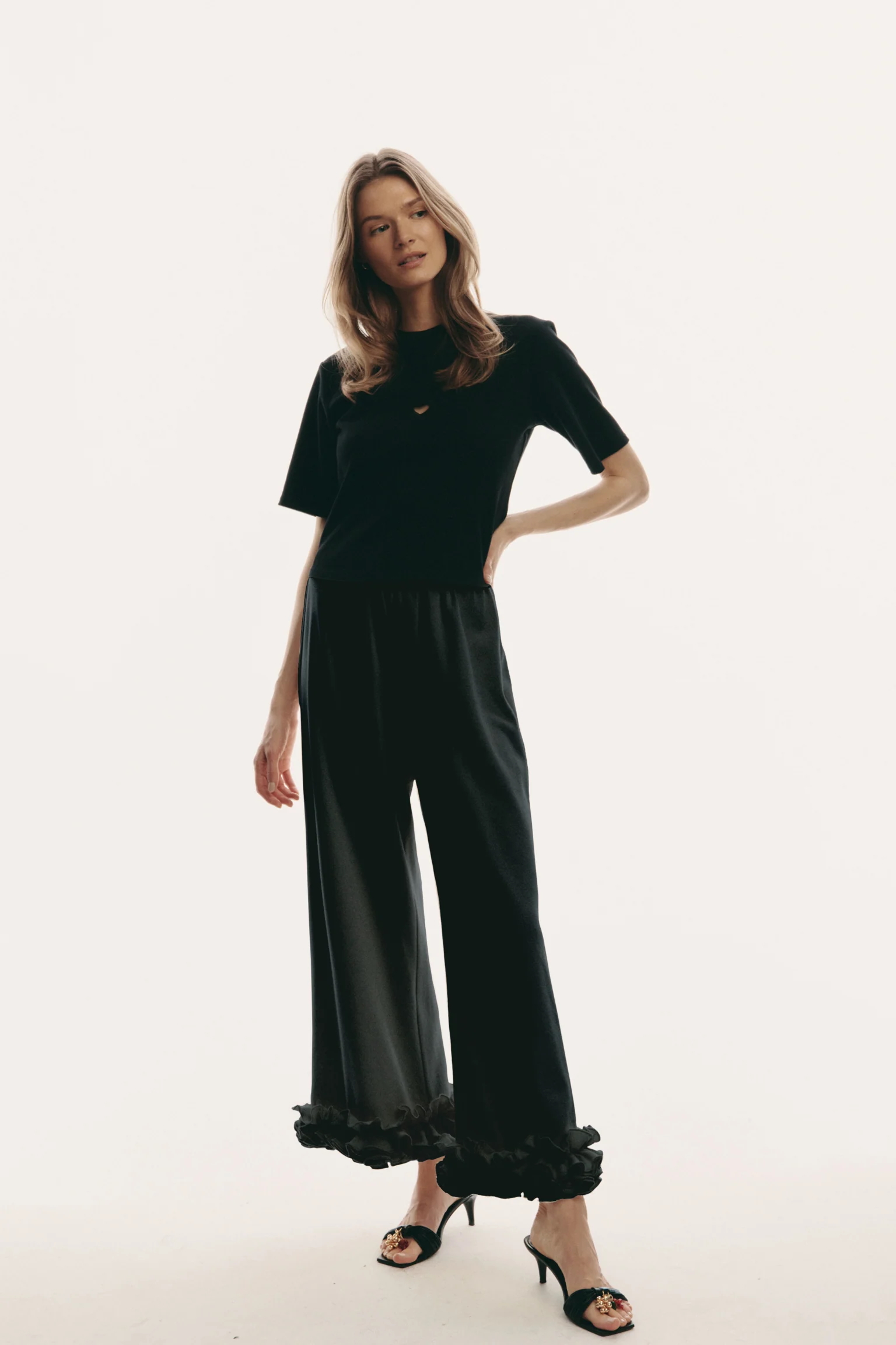 Modelka w czarnych spodniach z falbanką Le Petit Trou, prostym topie i eleganckich szpilkach.