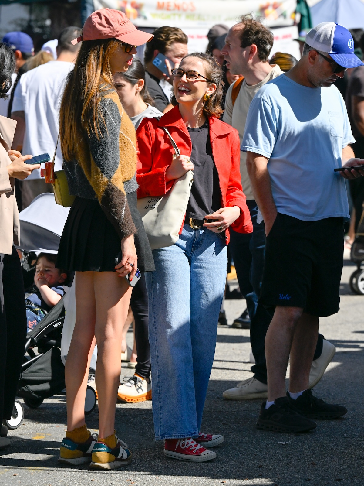 Aktorka Zoey Deutch w jasnych jeansach i czerwonych trampkach uśmiecha się do znajomej w tłumie na ulicy.