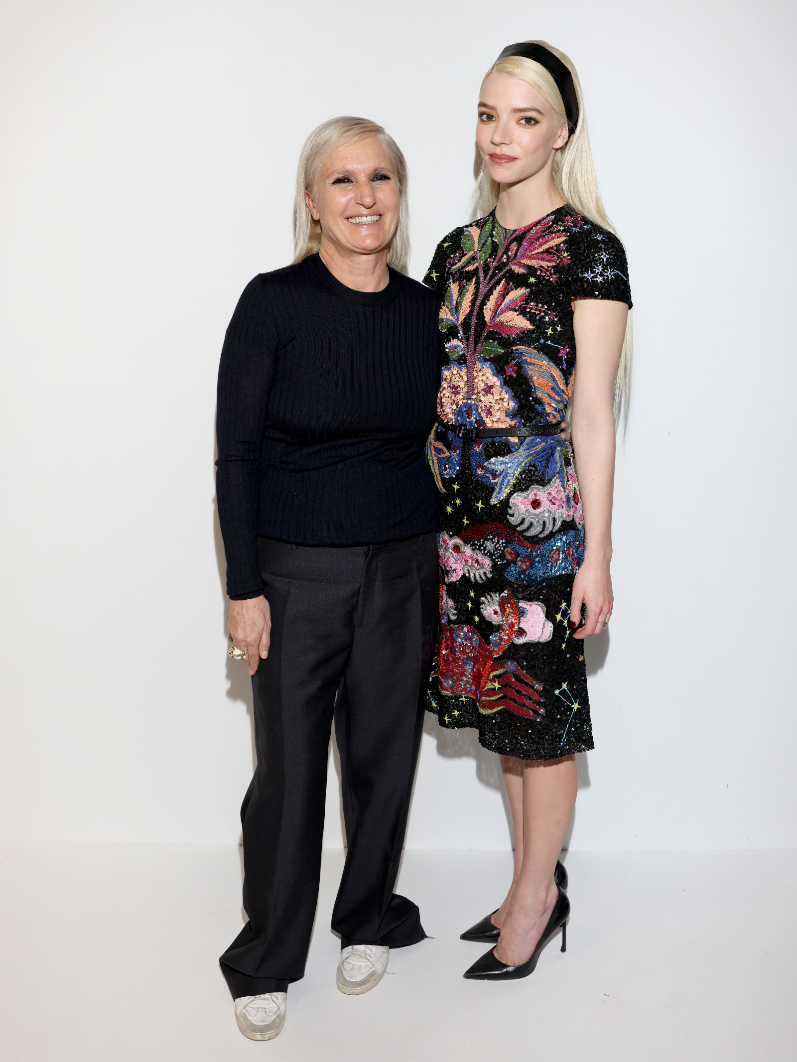 Maria Grazia Chiuri i Anya Taylor-Joy pozują razem, ubrane w eleganckie stylizacje, na neutralnym tle.