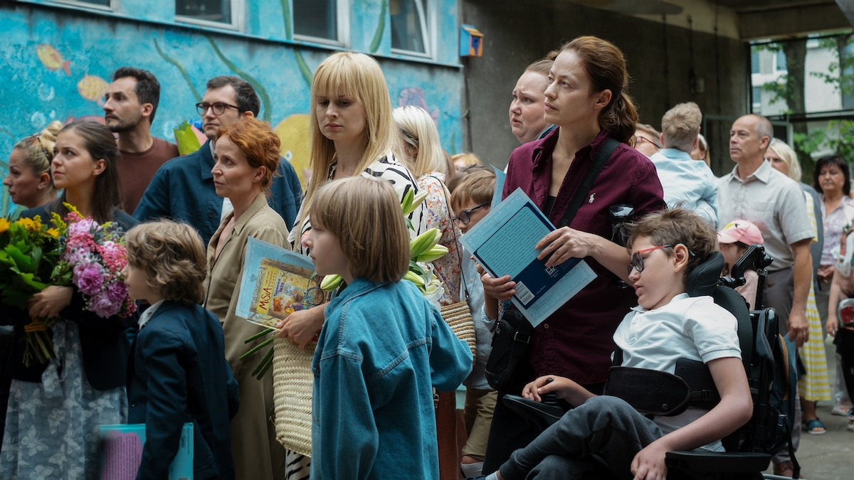 Kadr z polskiego serialu oryginalnego Netflix „Matki pingwinów”, przedstawiający grupę ludzi z kwiatami i dokumentami.