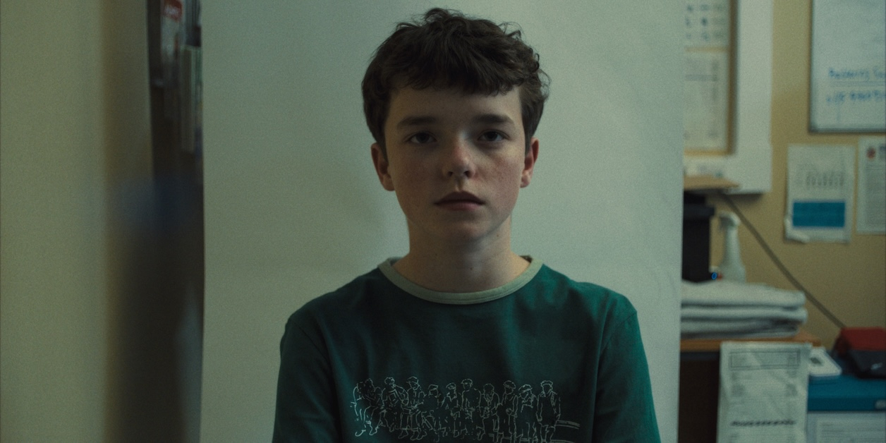 Owen Cooper z serialu Netflix „Dojrzewanie”, z wyrazem twarzy oddającym skupienie i refleksję, w tle minimalistyczne pomieszczenie.