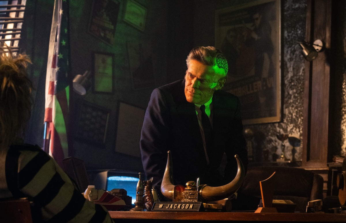 Willem Dafoe w filmie "Beetlejuice Beetlejuice", w stylowym biurze, z różnorodnymi rekwizytami w tle.