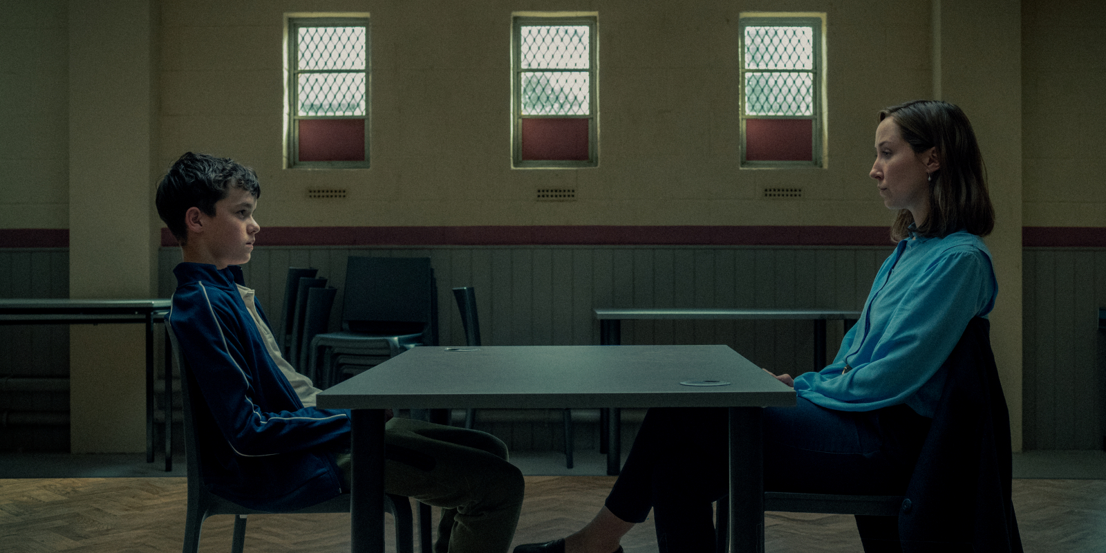 Owen Cooper i Erin Doherty siedzą naprzeciw siebie przy stole w szarym pomieszczeniu. Serial Netflix „Dojrzewanie”.
