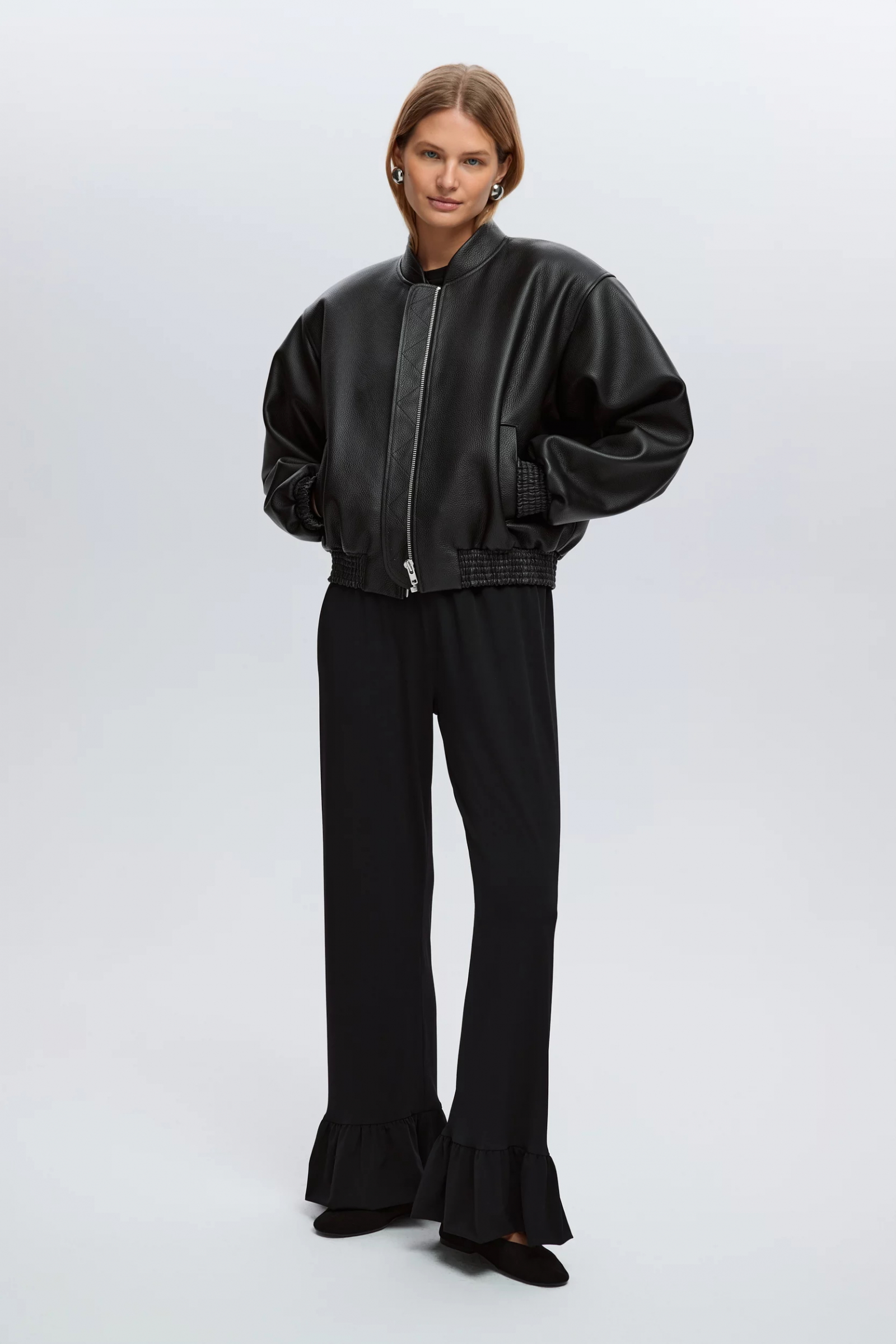 Kobieta w czarnej kurtce bomber, nosi czarne spodnie z falbanką Lui Store oraz czarne buty.