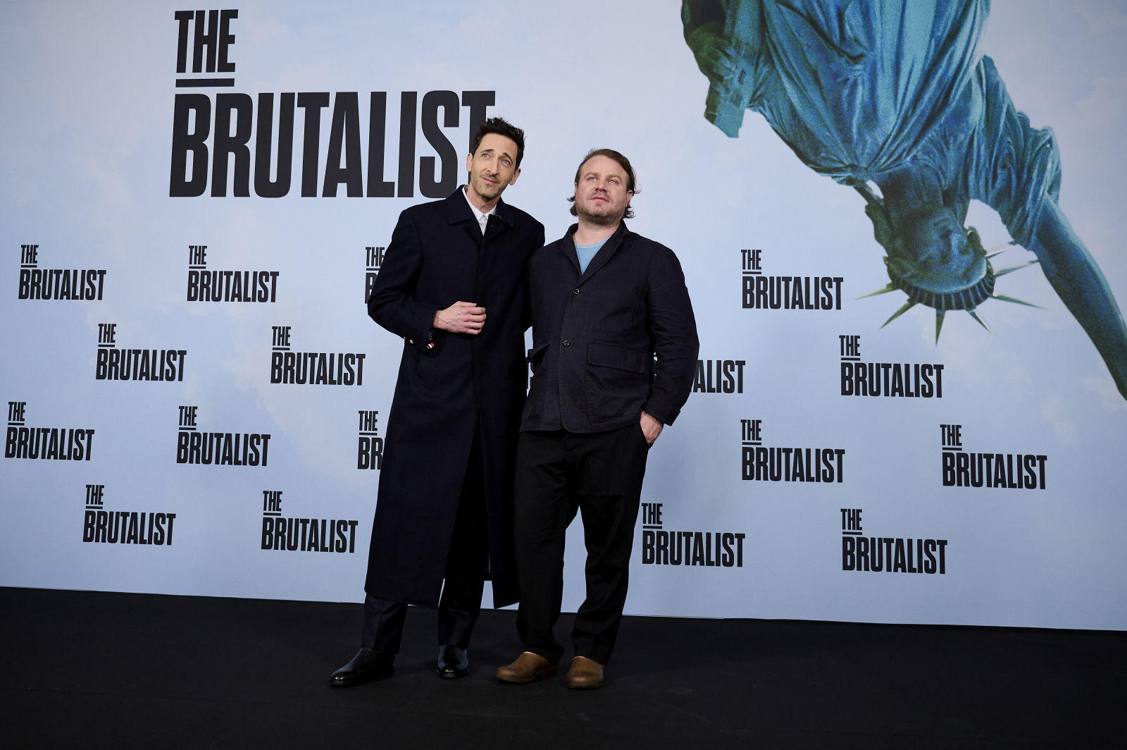 Adrien Brody i Brady Corbet na premierze filmu "The Brutalist", w tle Statua Wolności oraz logo filmu.