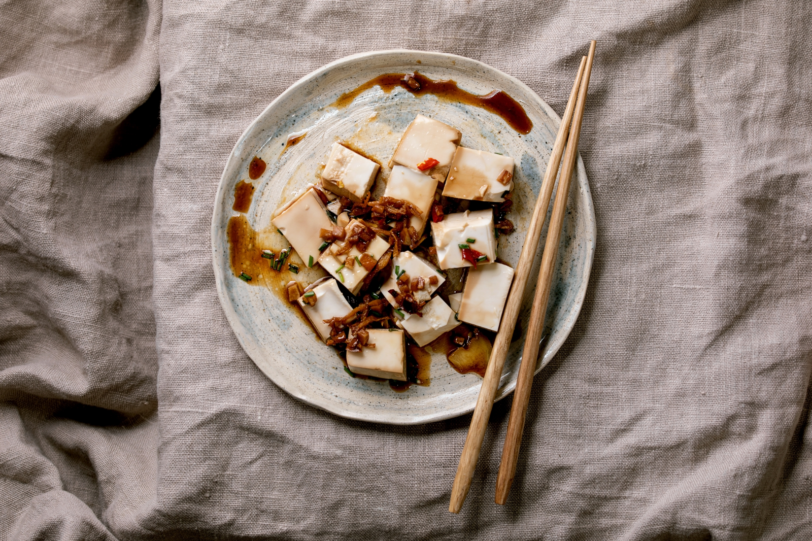 Talerz z pokrojonym tofu, posypanym szczypiorkiem i sosem, obok pałeczki na lnianym tle.