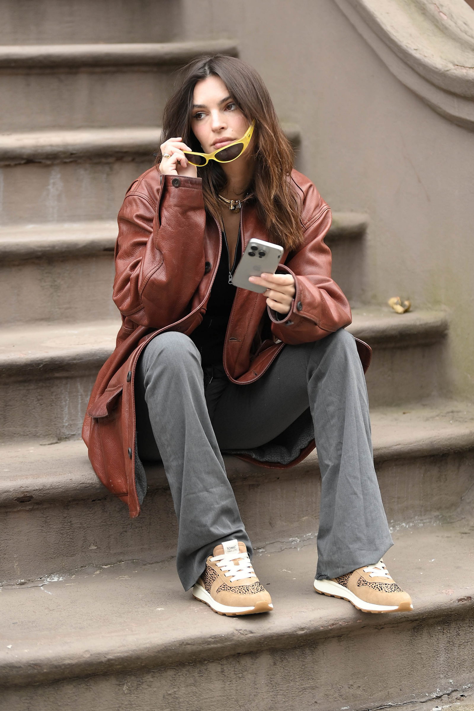Emily Ratajkowski w brązowej skórzanej kurtce i sneakersach w panterkę TOMS siedzi na schodach, trzymając telefon i okulary przeciwsłoneczne.