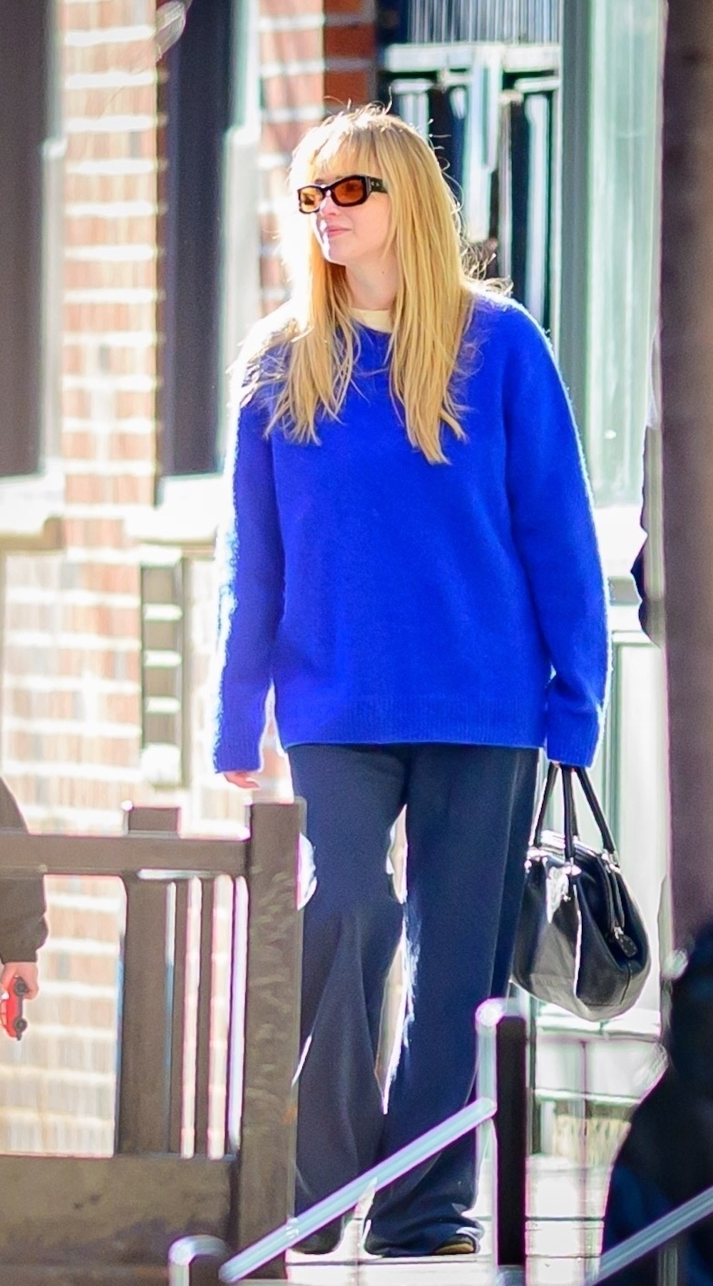 Jennifer Lawrence z blond grzywką i długimi włosami, ma na sobie intensywnie błękitny sweter marki The Row, niebieskie spodnie, czarną skórzaną torebkę i okulary przeciwsłoneczne z pomarańczowymi szkłami.