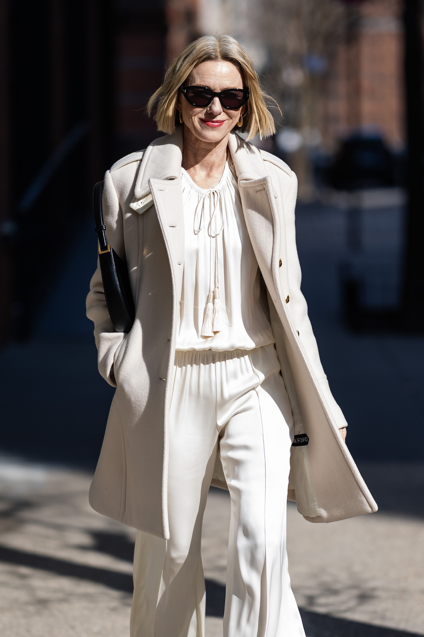 Naomi Watts ma na sobie biały płaszcz, białą koszulę ze szureczkami, białe spodnie z szeroką nogawką, czarną skórzaną torebkę i czarne okulary przeciwsłoneczne.