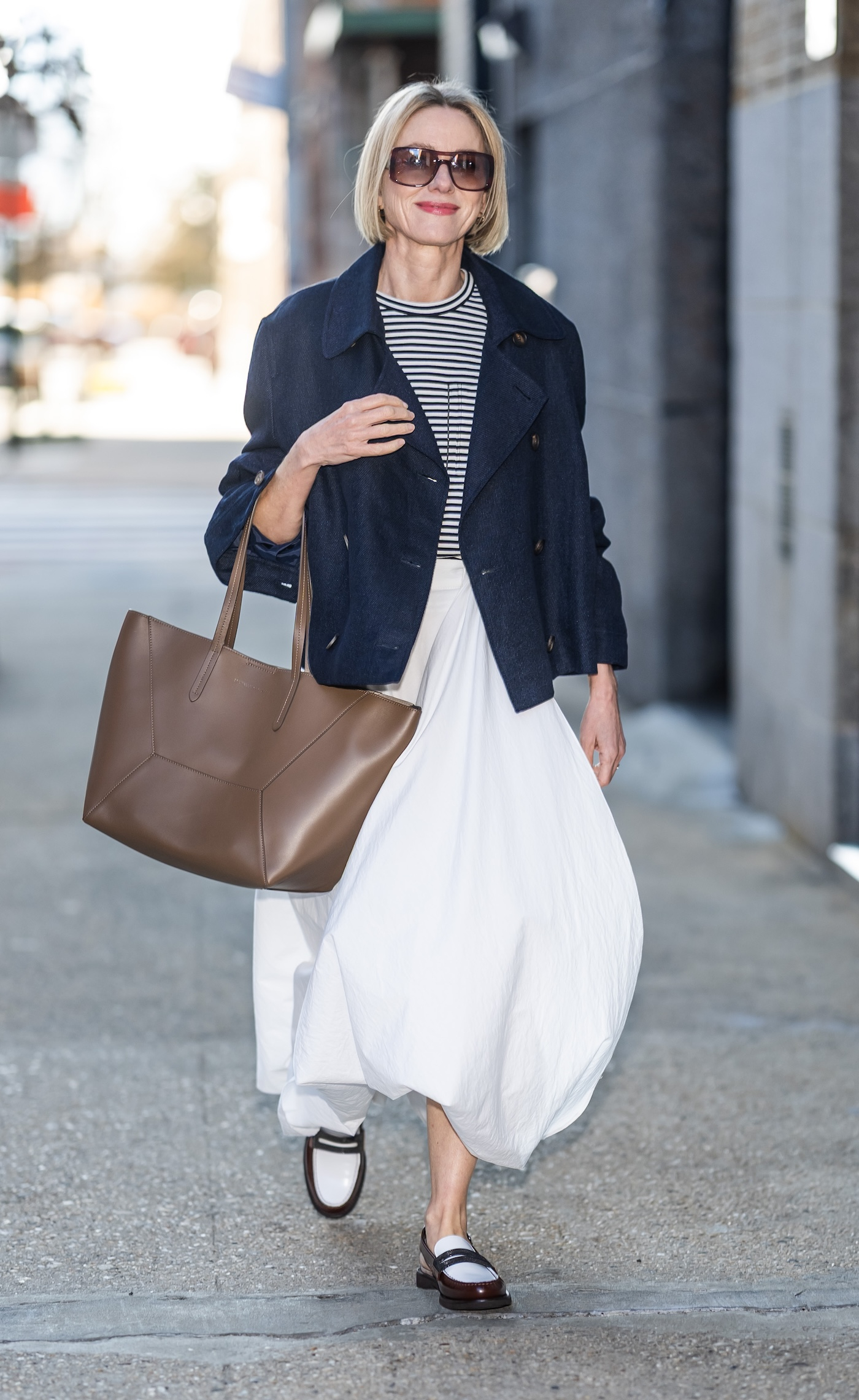 Naomi Watts ma na sobie bluzkę w paski, krótki trencz, rozkloszowaną białą spódnicę midaxi, skórzaną brązową torebkę i dwukolorowe mokasyny.