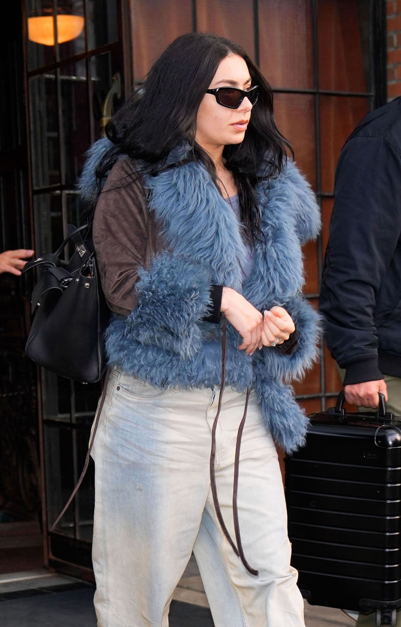 Charli XCX w czarnych okularach przeciwsłonecznych, jasnych baggy jeans i kurtce z niebieskim futerkiem.
