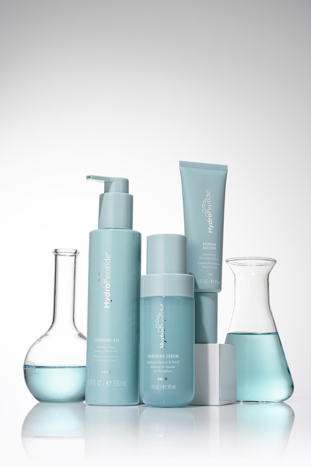 Produkty HydroPeptide w jasnoniebieskich, pastelowych opakowaniach: żel oczyszczający, serum łagodzące i budujący barierę, obok szklanych naczyń laboratoryjnych.