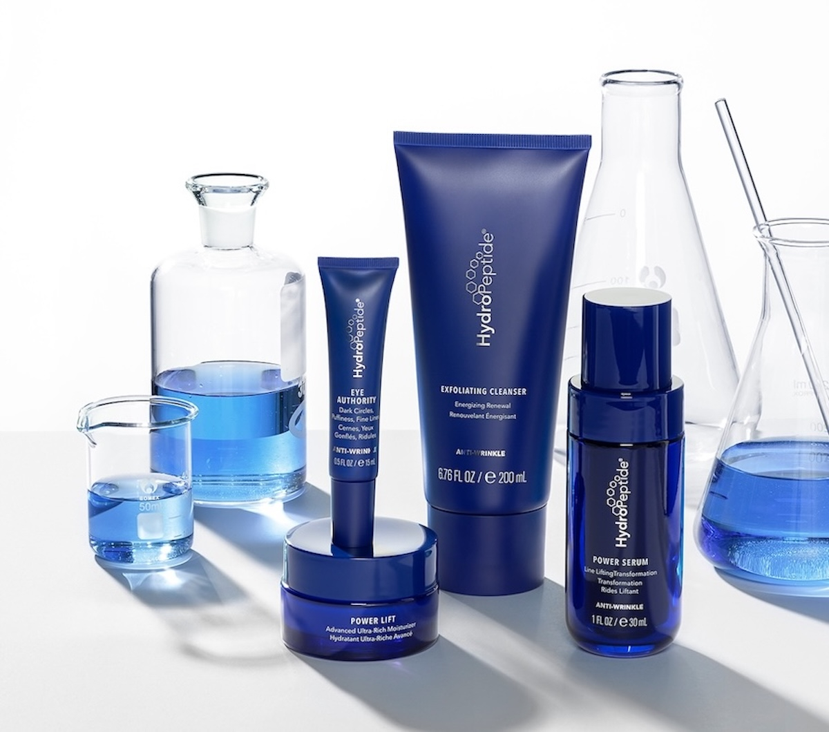 Na zdjęciu znajduje się linia produktów HydroPeptide do pielęgnacji skóry: oczyszczający żel, serum, krem w otoczeniu szkła laboratoryjnego.