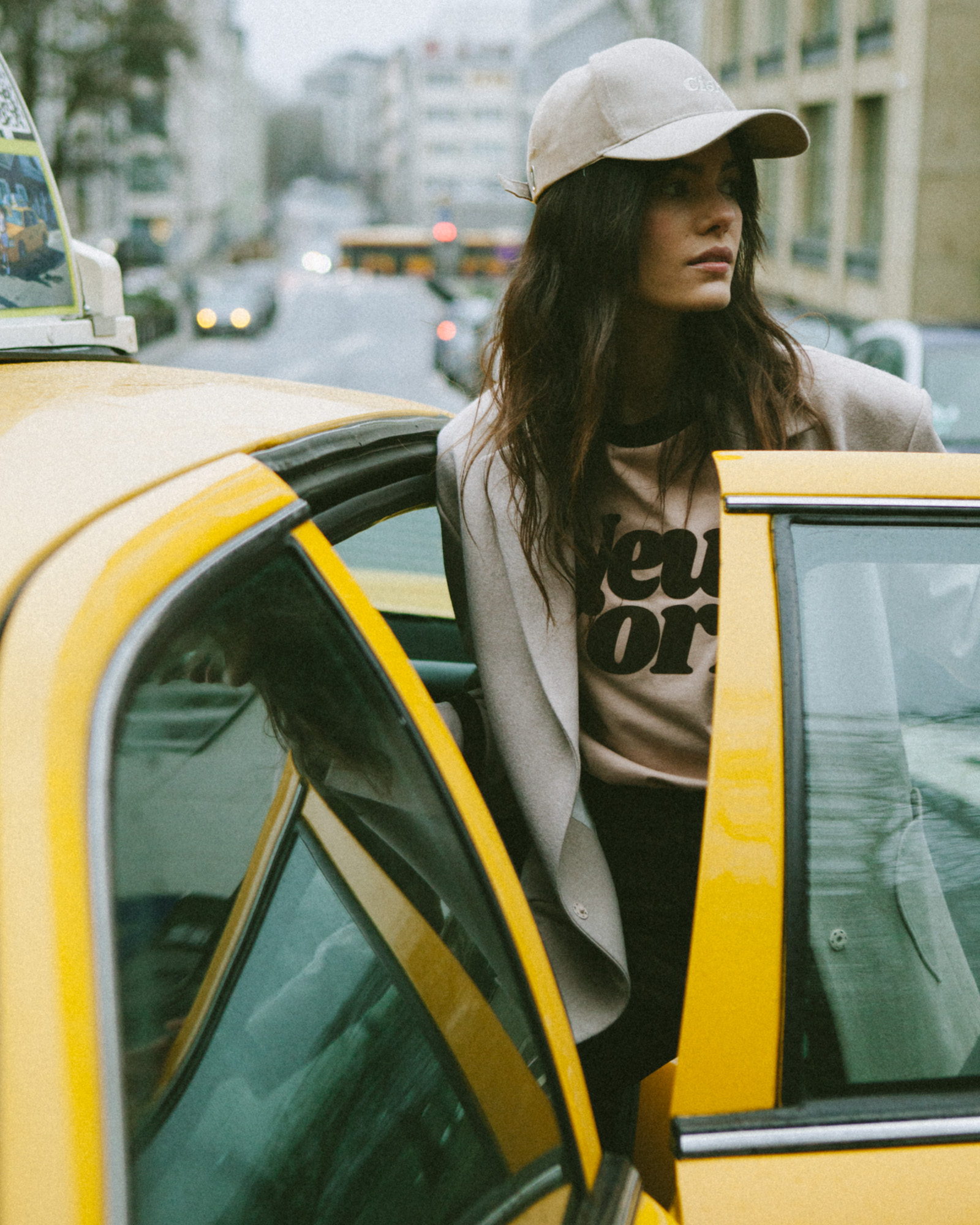 Kobieta w czapce wsiada do żółtego taxi, ubrana w bluzę She Is Sunday z napisem "New York" i szary żakiet. Tło miasta w mglisty dzień.