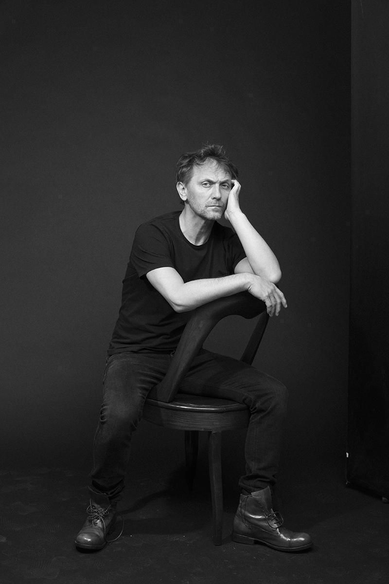 Andrzej Chyra siedzący na krześle w czarnym t-shircie i jeansach.