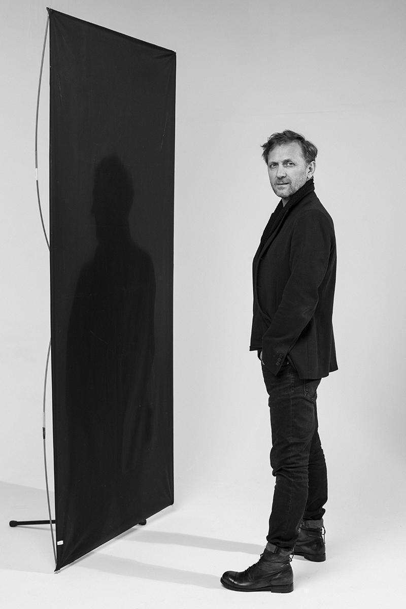 Andrzej Chyra stojący przed ścianą z czarnego materiału, na której widać jego cień.