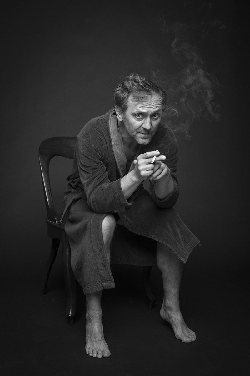 Andrzej Chyra siedzi na krześle w szlafroku i pali papierosa.