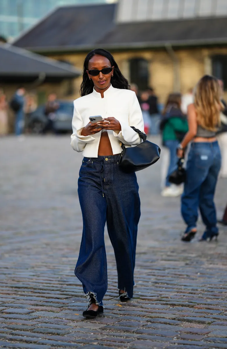 Kobieta w białej kurtce i damskich jeansach spaceruje w eleganckich butach, trzymając telefon w ręku.