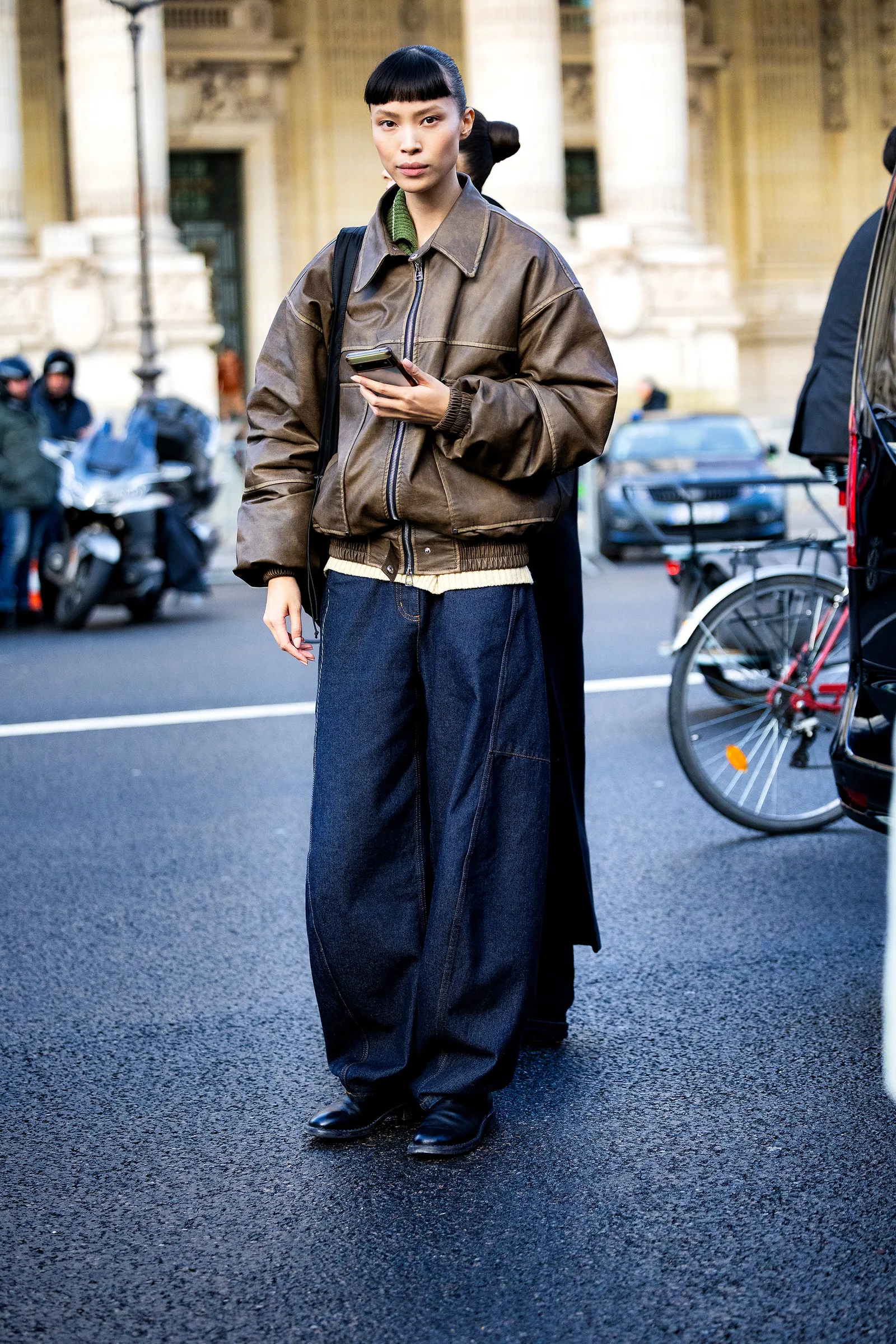 Osoba w eleganckich butach oraz damskich jeansach, trzymająca telefon, stoi na ulicy. W tle widać ruch uliczny.