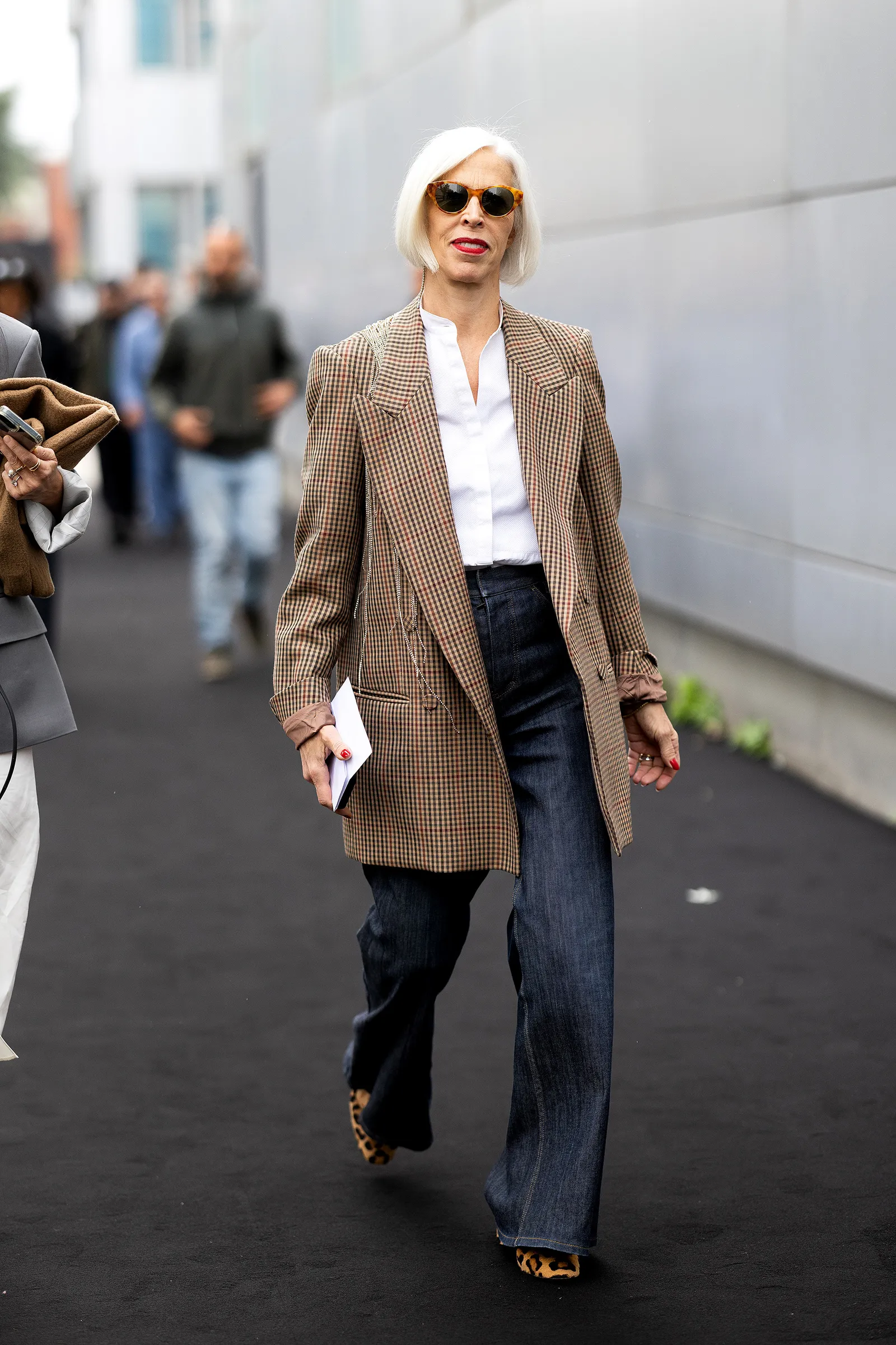 Kobieta w eleganckim płaszczu, nosząca jeansy damskie i eleganckie buty, spaceruje po ulicy w miejskim otoczeniu.