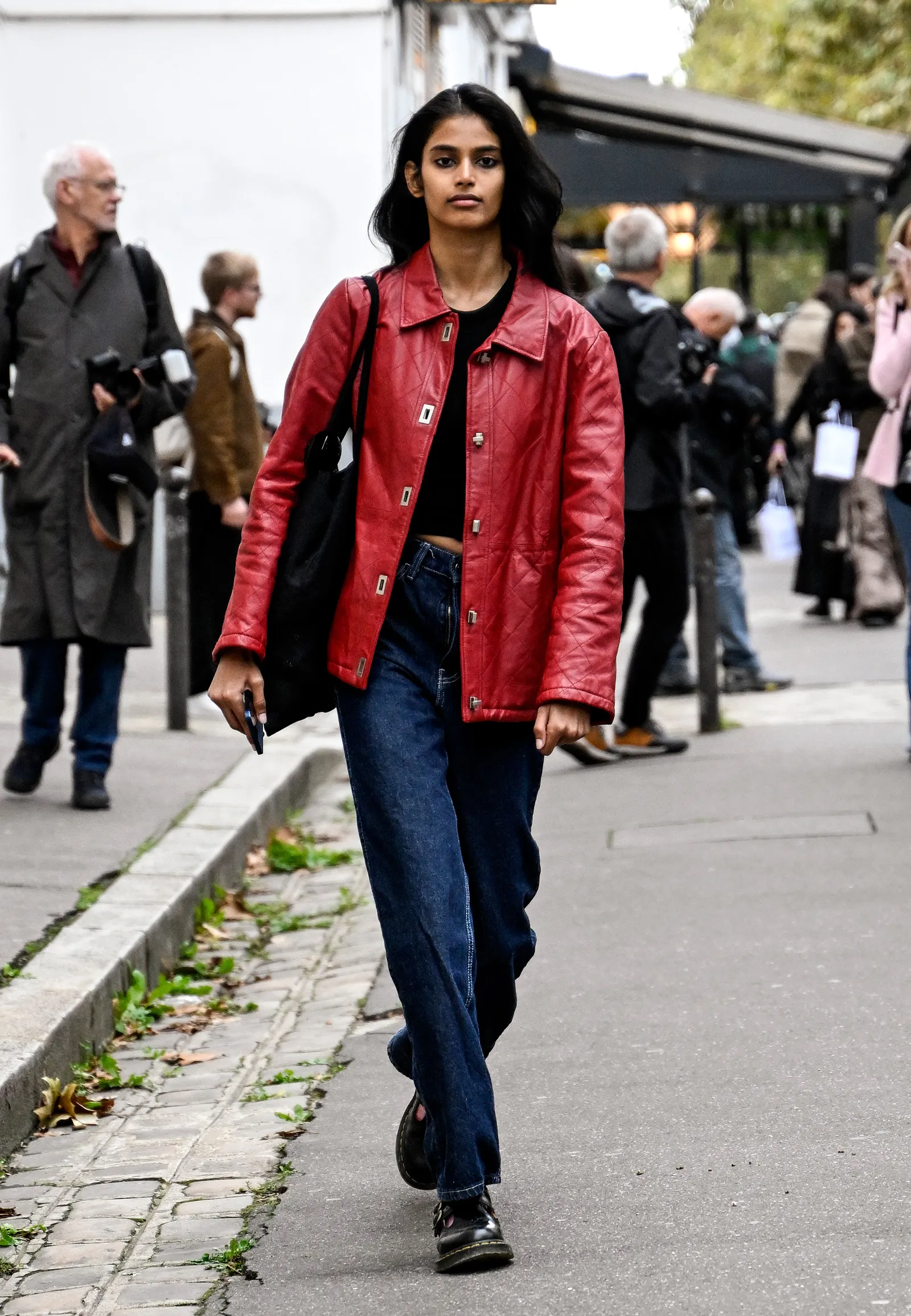 Kobieta w czerwonej kurtce, jeansy damskie i eleganckie buty idzie po chodniku, w tle inne osoby.