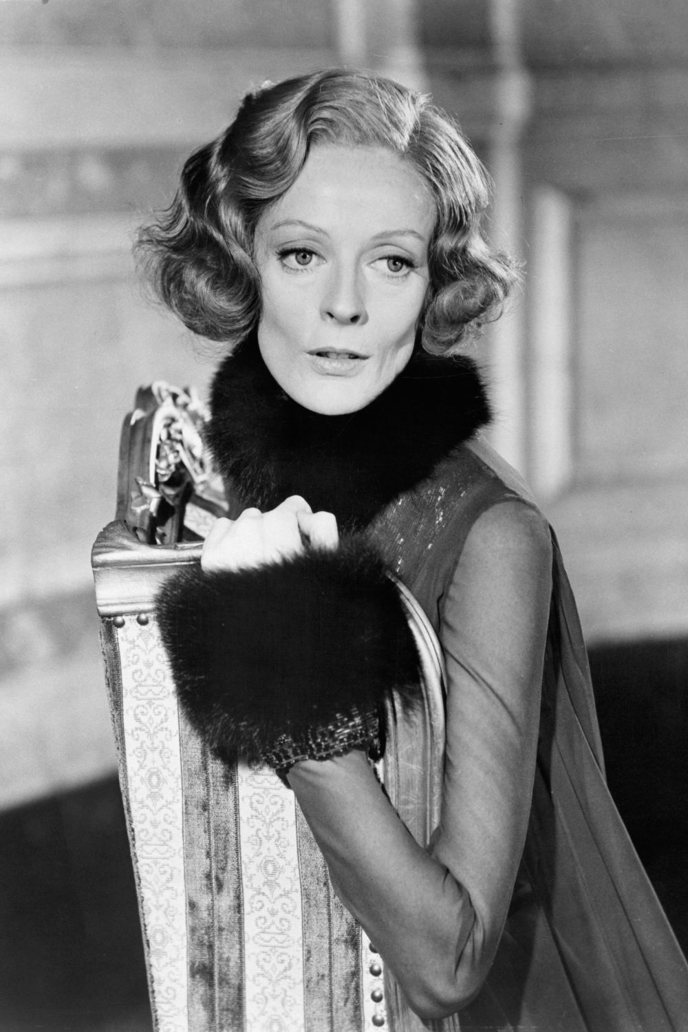 Maggie Smith w eleganckiej kreacji, trzymająca walizkę z futrzanym wykończeniem, patrzy z wyrazem refleksji.